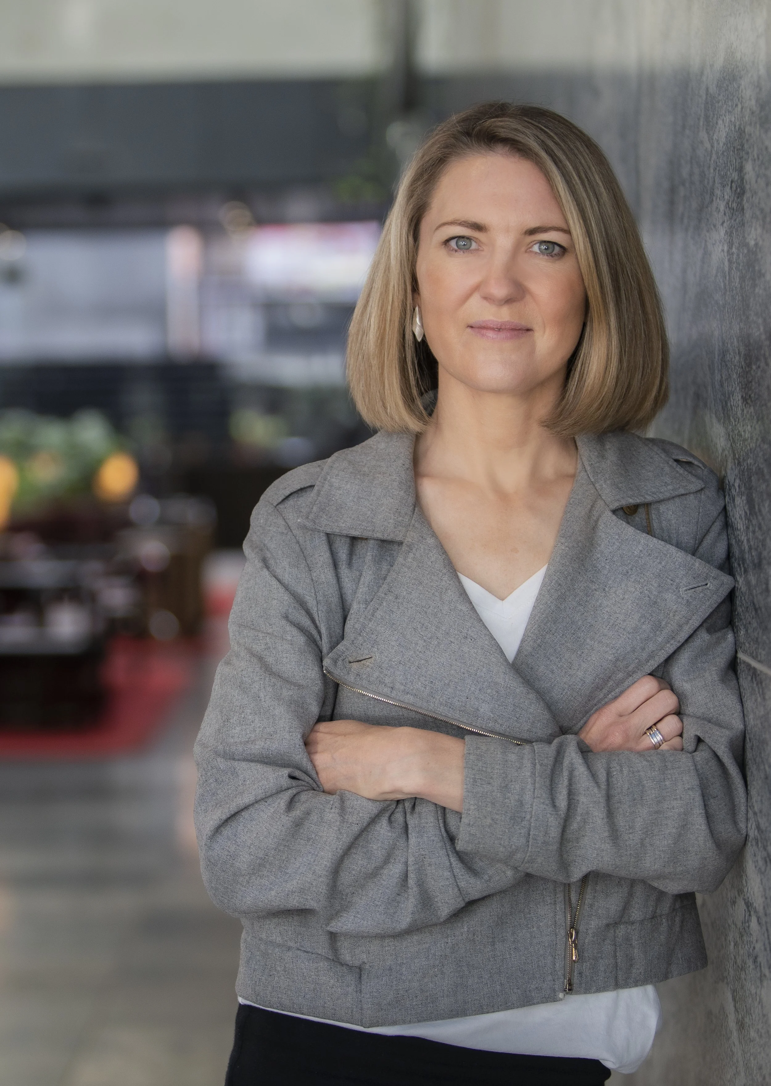 A woman with shoulder-length blonde hair and blue eyes leaning against a gray wall, crossing her arms and looking at the camera. She is wearing a gray jacket and a white shirt underneath, with a ring on her finger. The background is blurry with some plants and parking structures.