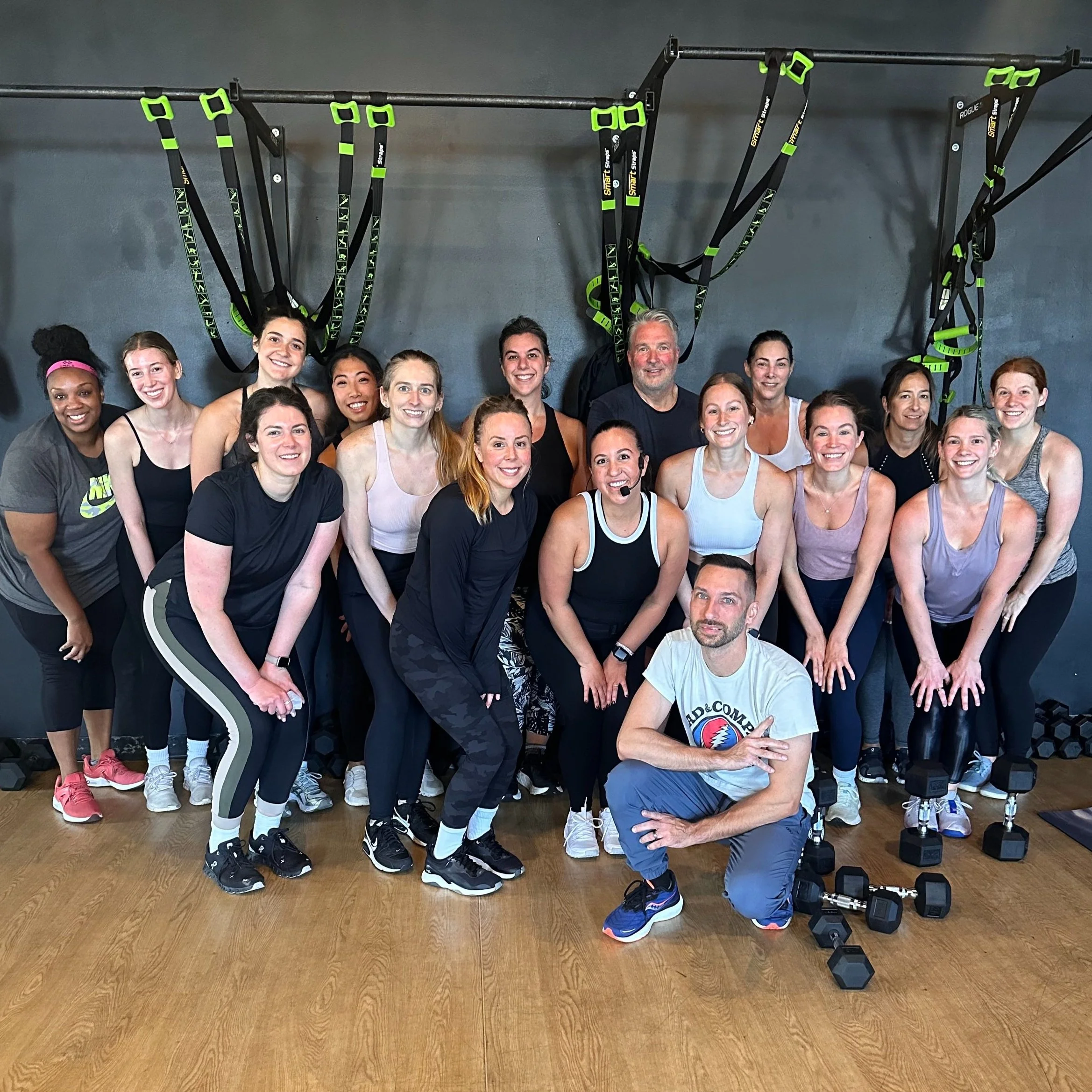 Group of people in a fitness class posing for a photo, with exercise straps and dumbbells in the background.