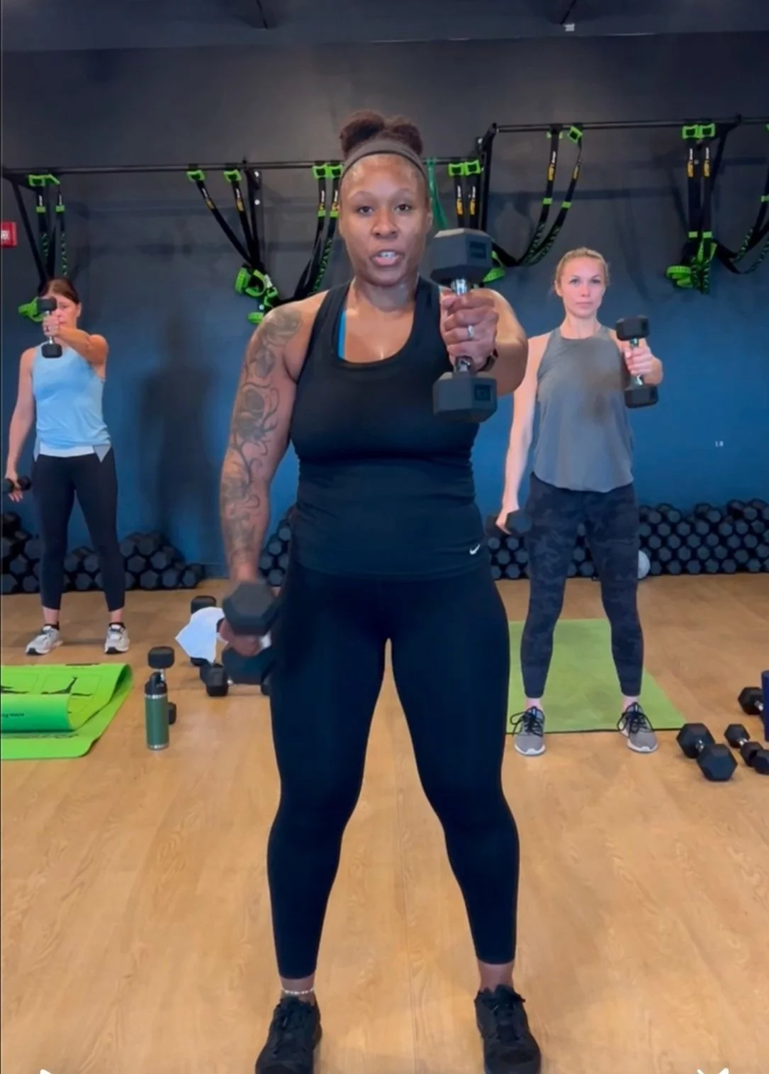 Three women exercising with dumbbells in a gym, standing on a wooden floor with exercise mats and weights around them, and fitness equipment hanging on the wall behind them.