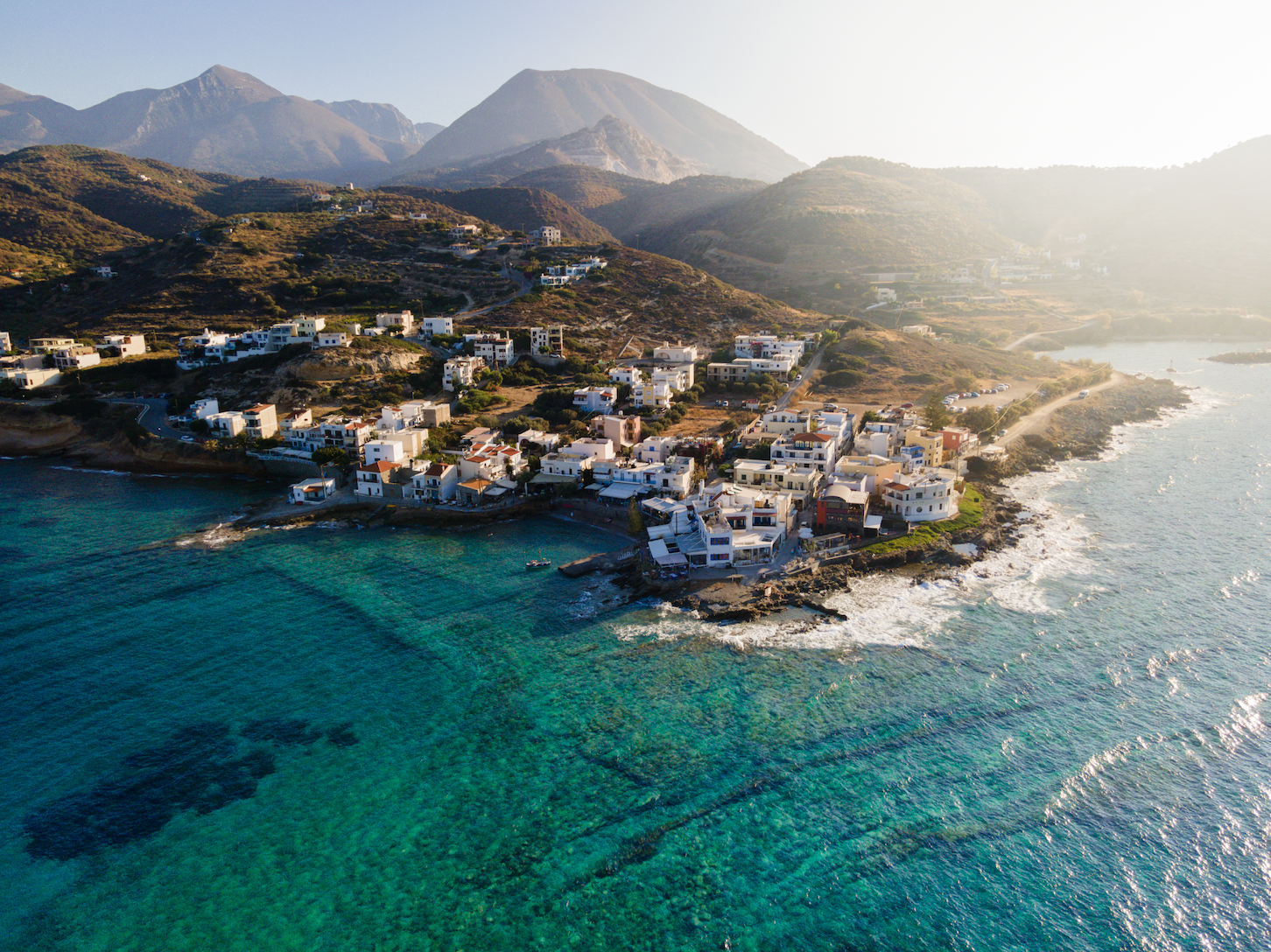 Aerial view of a coastal town with white buildings along the shoreline, surrounded by mountains, with clear blue water and a boat near the shore.