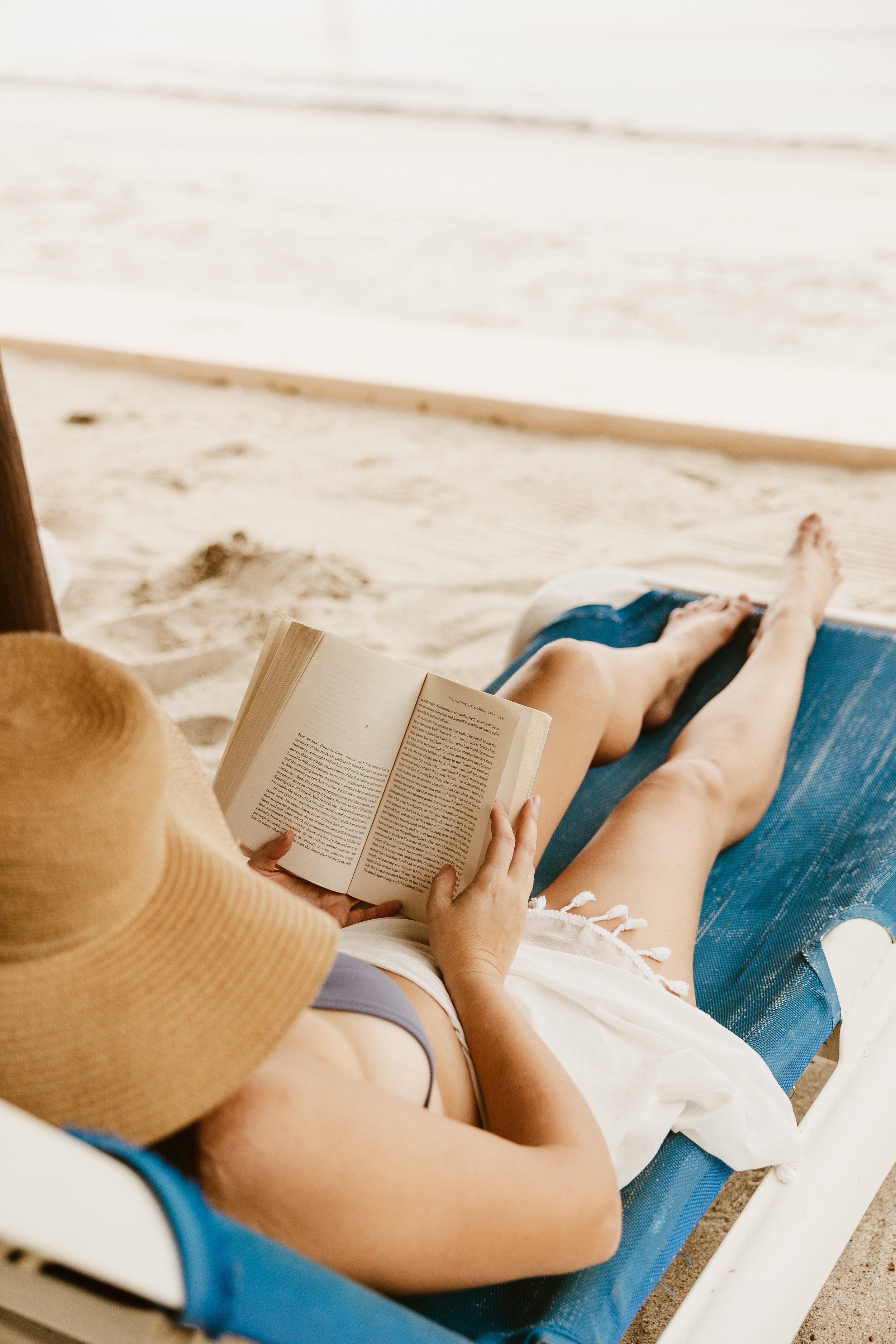 Person in a yellow hat, tan top, and gray shorts sitting on a blue lounge chair on a sandy beach, reading a book.