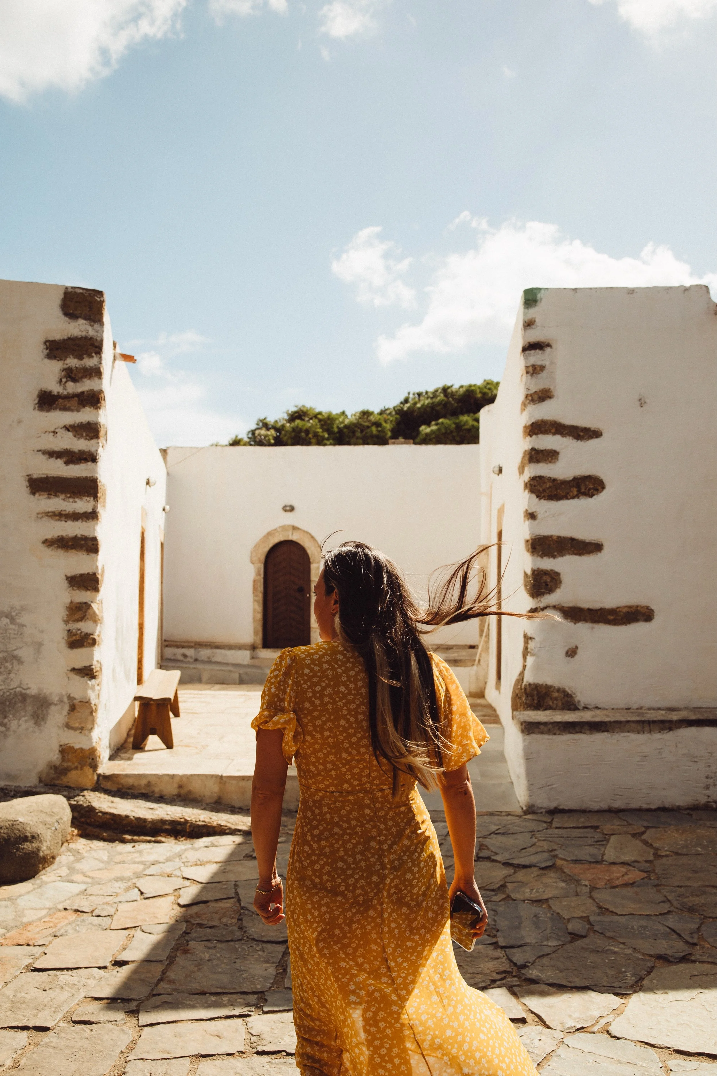 A woman with long hair wearing a yellow dress with white floral pattern walking through a sunny courtyard with white walls and stone flooring.