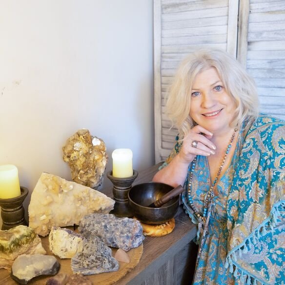 A woman in a blue patterned robe stands next to a table with assorted crystals and stones, two lit candles, and a singing bowl.