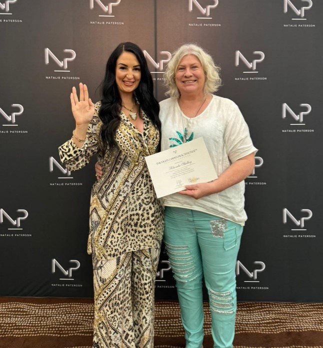 Two people Rhonda and Natalie Paterson posing in front of a backdrop with "Natalie Paterson" and "NP" logos. One person is holding a certificate and the other is waving.