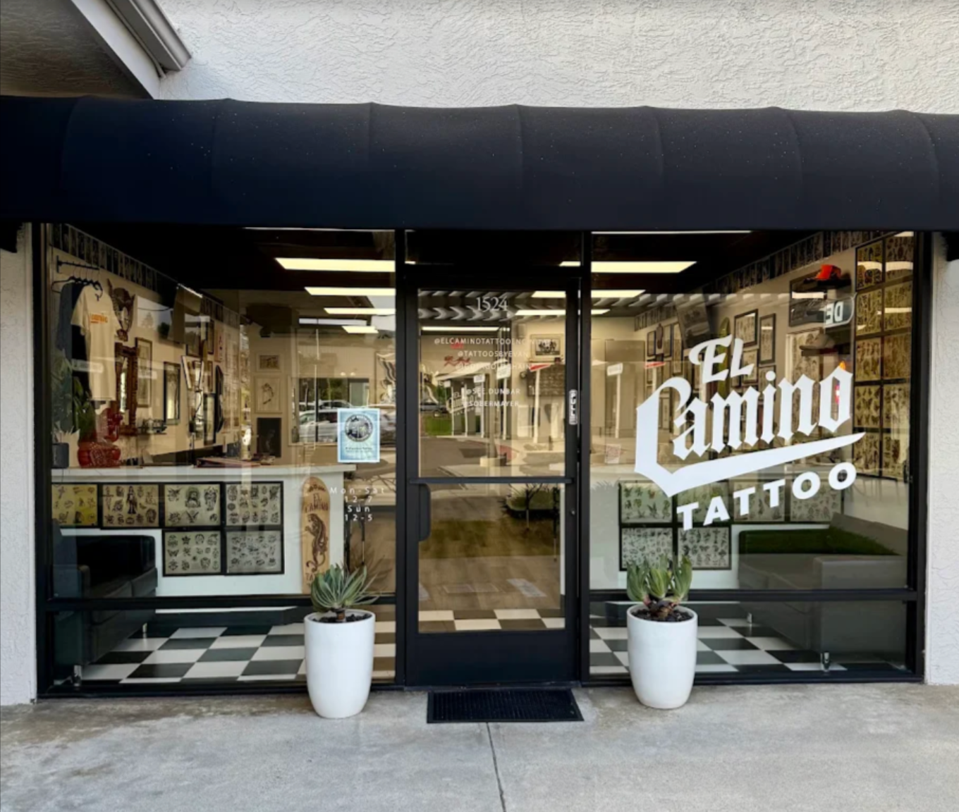 The storefront of a tattoo shop named El Camino Tattoo, with large glass windows and doors, black awning, and two white potted plants outside. Inside, framed artwork and tattoo designs are visible.