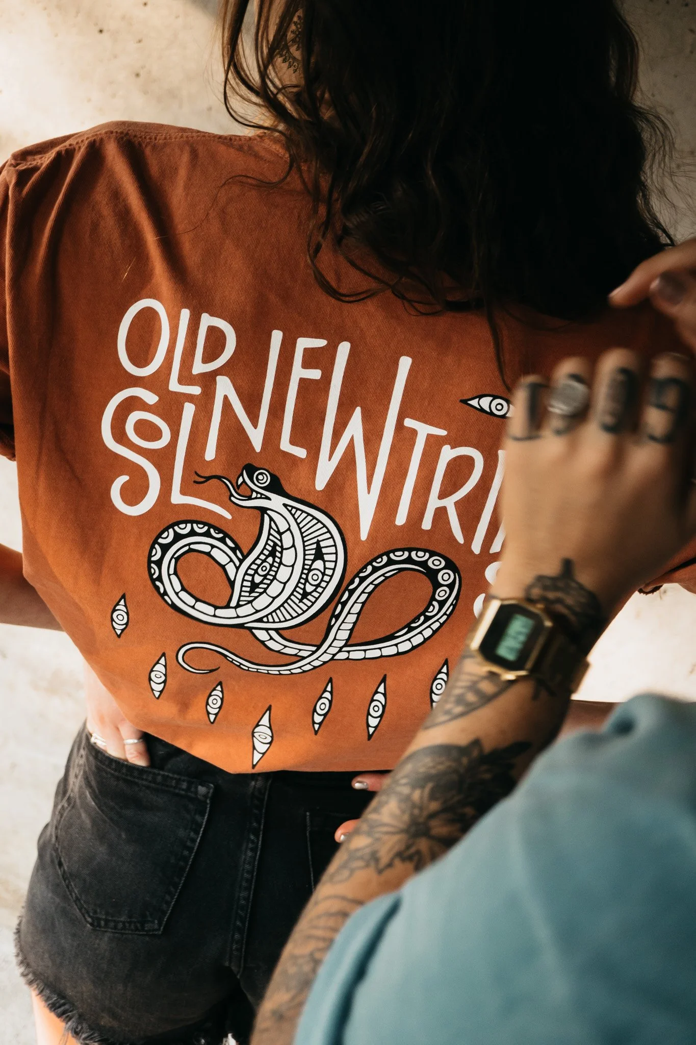 A person wearing an adobe colored Old Sol New Tricks graphic t-shirt with a snake illustration, and black shorts. The person has long dark hair. Another person with tattoos on their forearm is touching their shoulder.