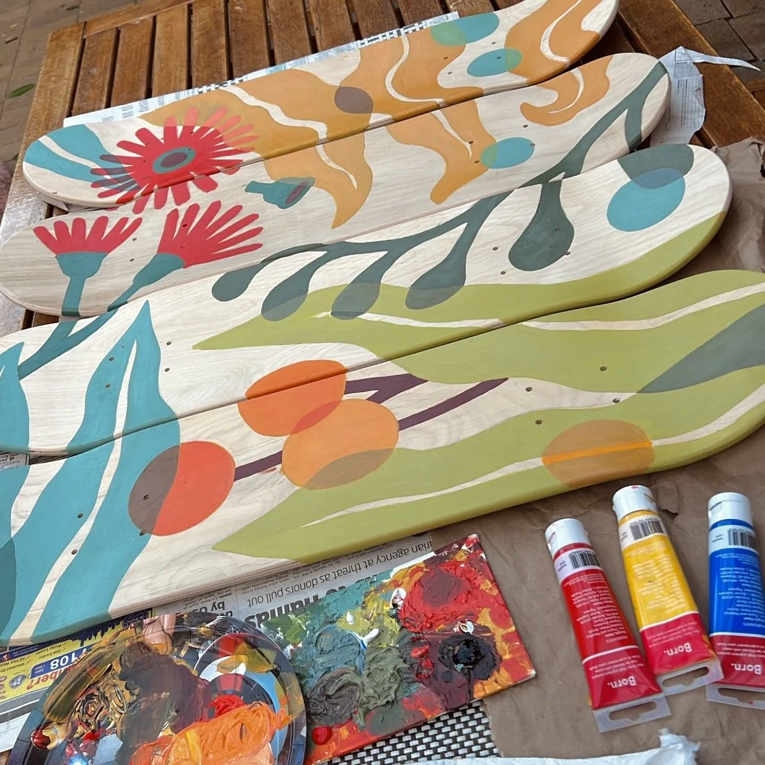 Painted wooden skateboards with colorful floral and botanical designs, placed on a wooden surface with paint tubes and palette nearby.