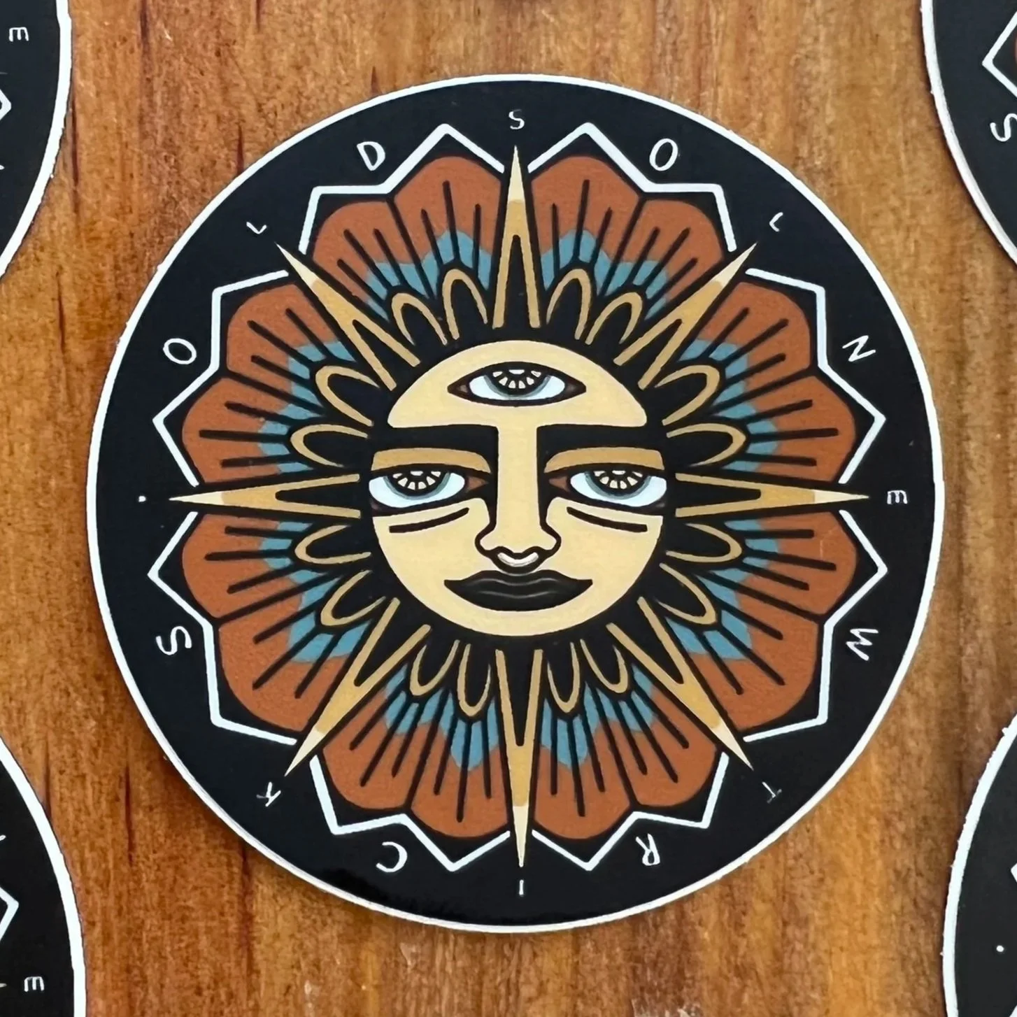 A round decorative Old Sol New Tricks brand sticker featuring a stylized face with three eyes, dark lips, and a sunburst design with colored segments around the face, on a wooden background.