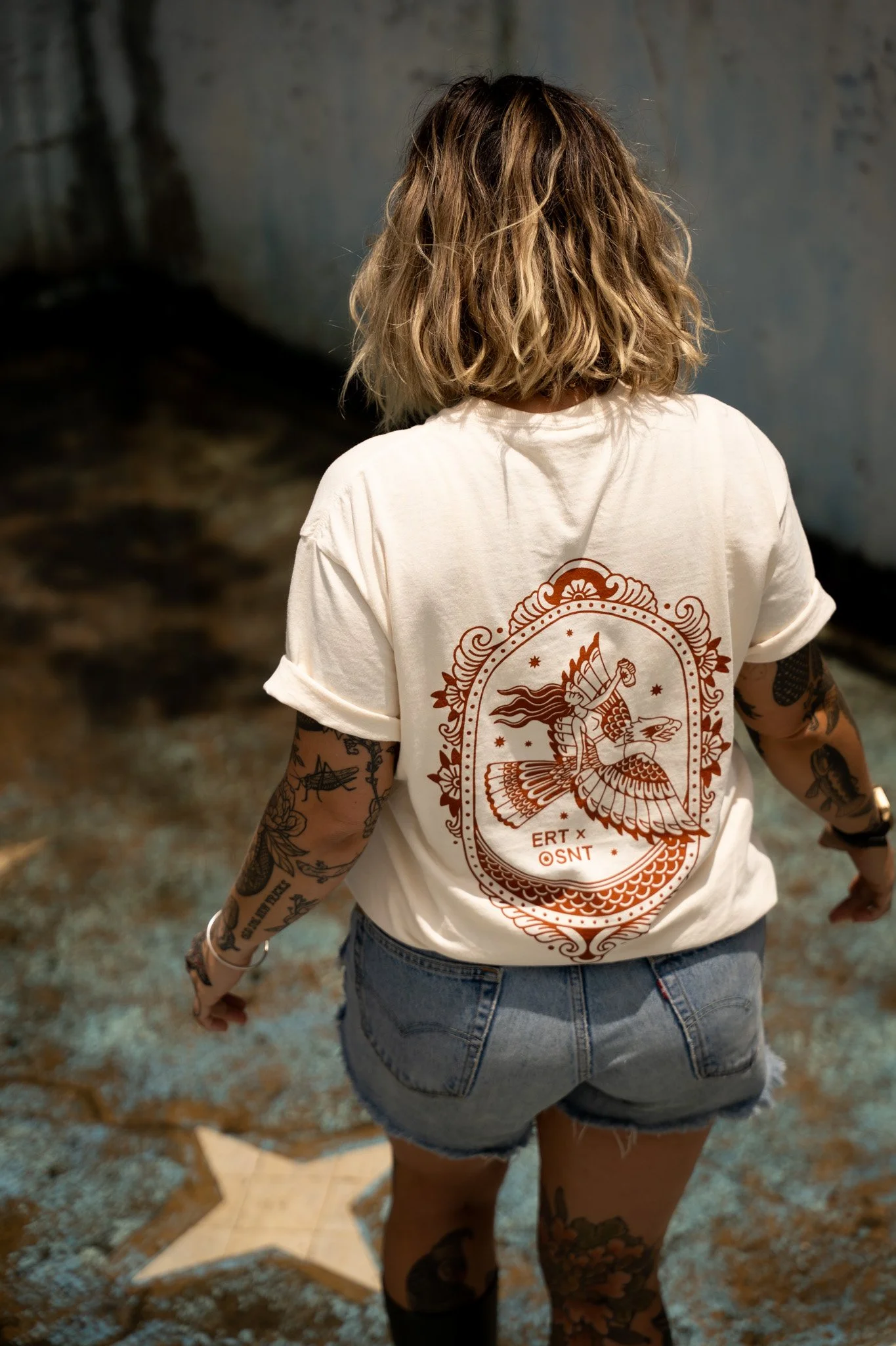 Cat James with tattoos on arms and legs, wearing a white t-shirt with a graphic design collaboration on the back, and denim shorts, standing in an empty skate pool.