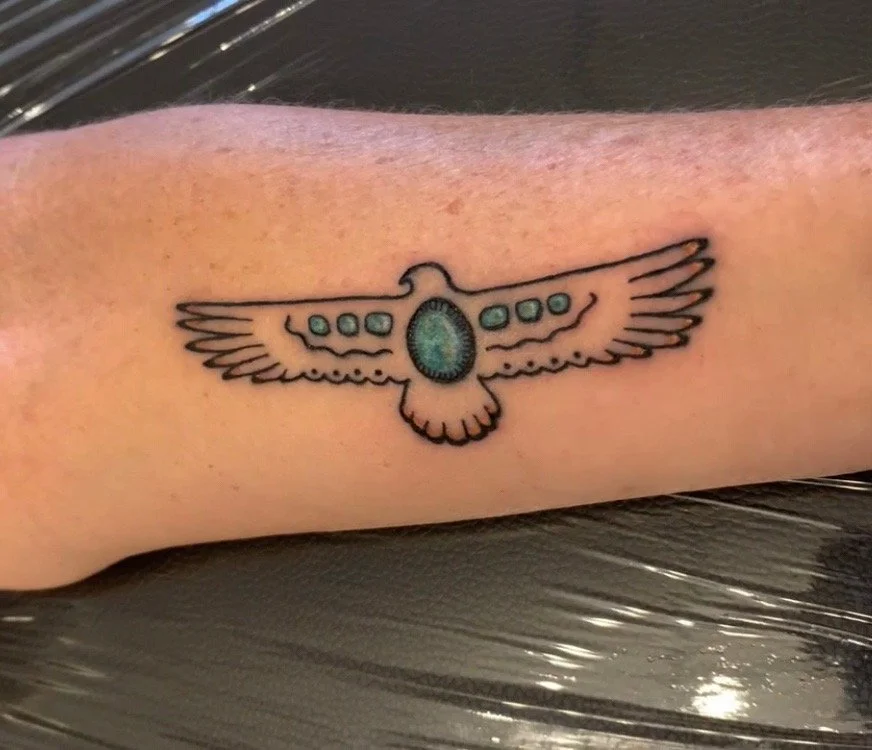Tattoo of a bird with outstretched wings, featuring a decorative turquoise and black gemstone in the center, on a person's forearm.