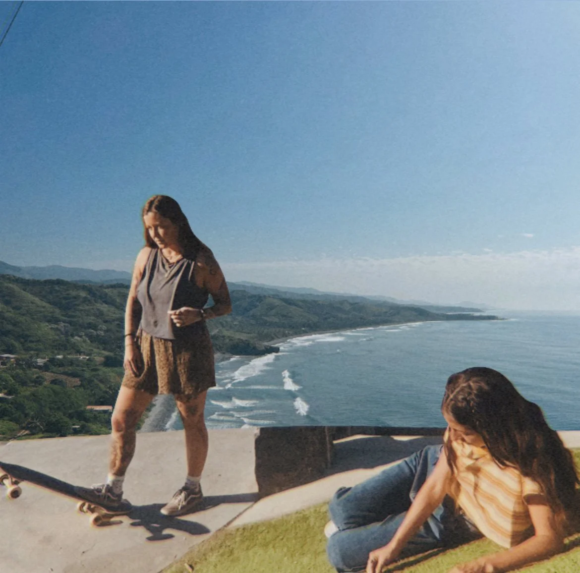 Two women on a grassy hill overlooking the ocean with waves and green mountains in the background. One is standing on a skateboard, and the other is sitting on the grass.