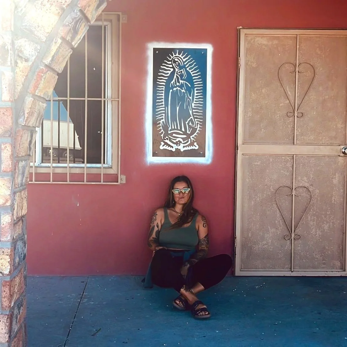 A woman with tattoos and sunglasses sitting cross-legged on the sidewalk with her back against a red wall, beside a large decorative image of Our Lady of Guadalupe, with an arched opening made of brick on the left and a metal door with heart-shaped patterns on the right.