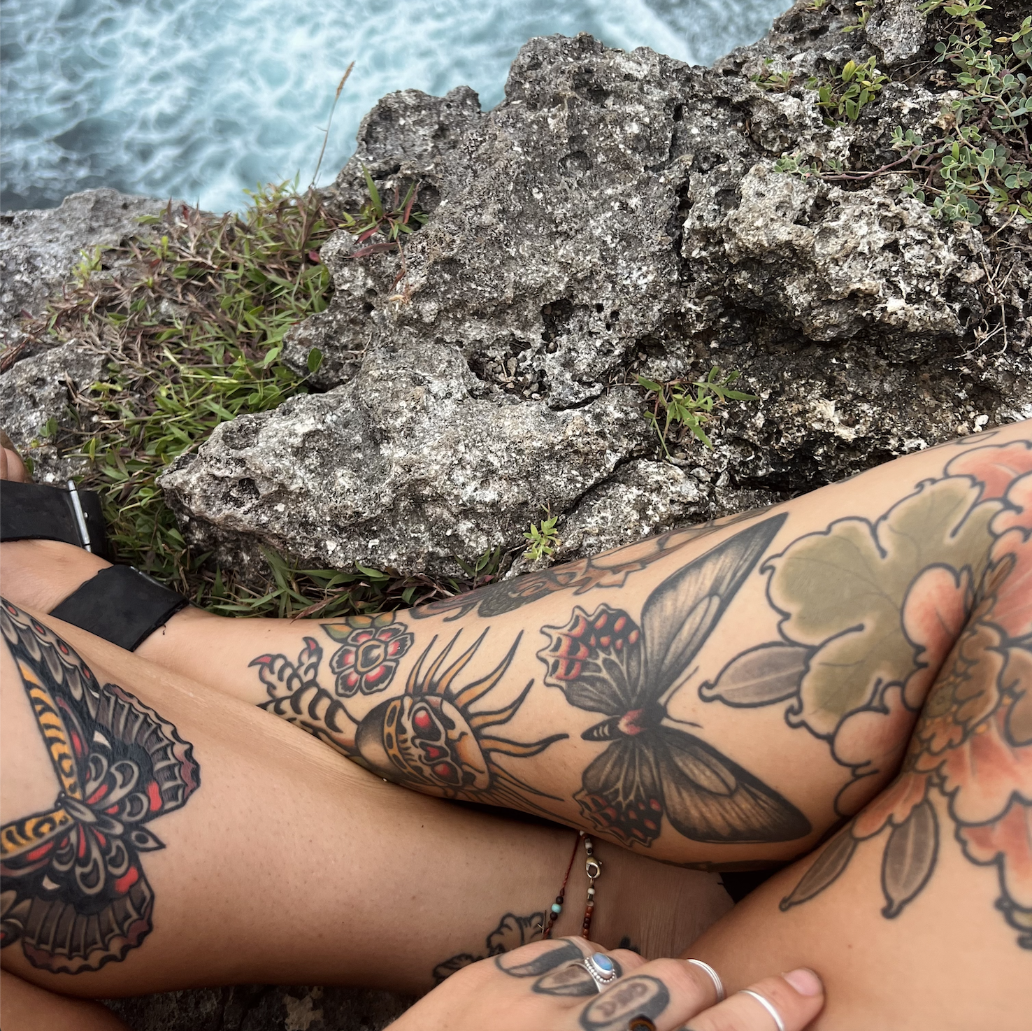 A person with tattoos on their arms and legs, wearing rings, lying near a rocky cliff inn Bali, Indonesia with green plants, overlooking a body of water.