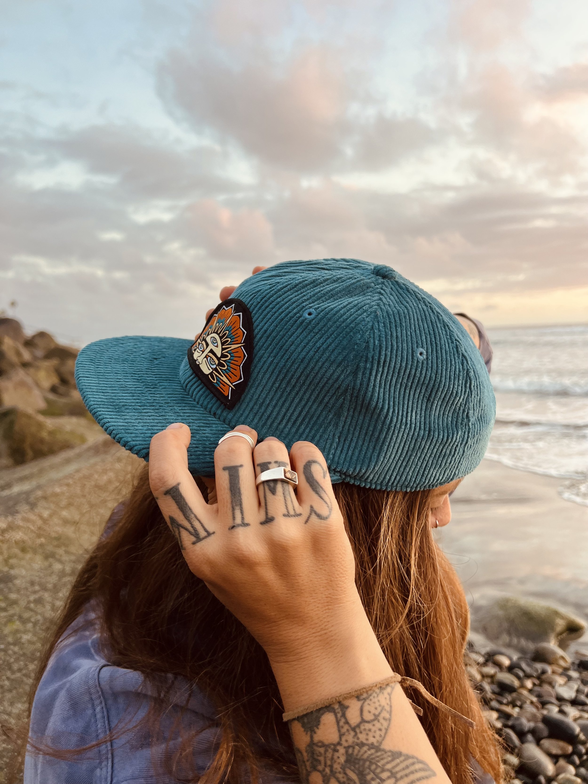 Person wearing a teal corduroy baseball cap with an old sol new tricks logo patch, holding their hand to the side of their face, at oceanside beach during sunset.
