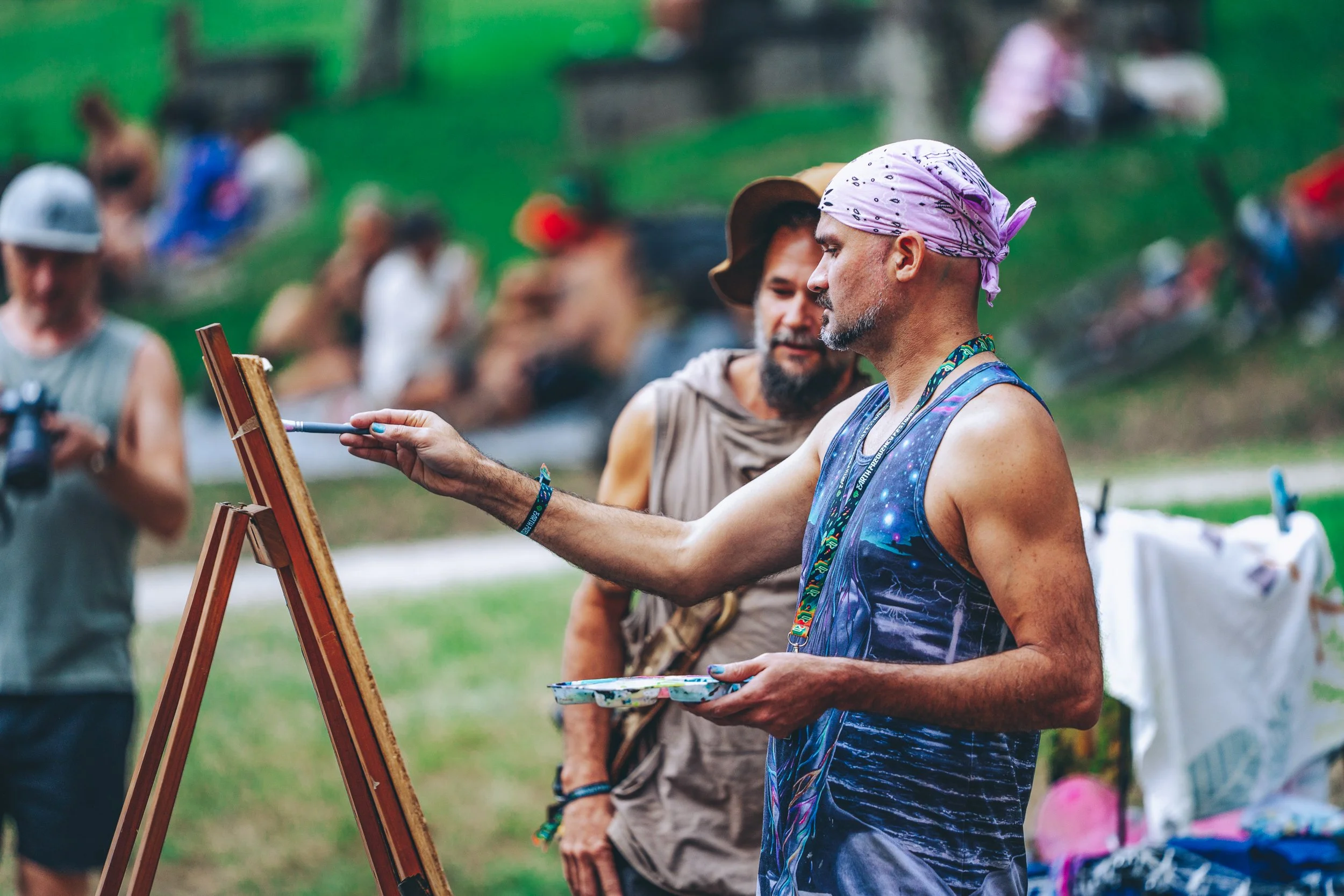 A man with a pink bandana and galaxy-themed tank top paints on a canvas outdoors as others watch on a grassy area.