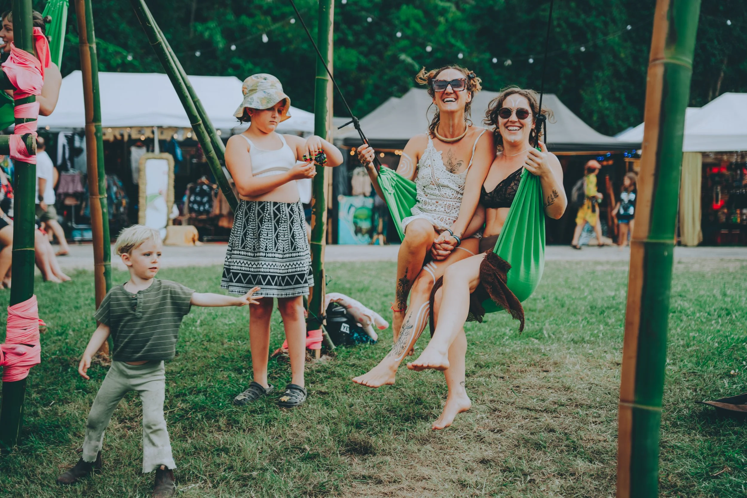 Two women sitting on a swing, smiling and enjoying a day outdoors at a park or festival, surrounded by children and tents in the background.