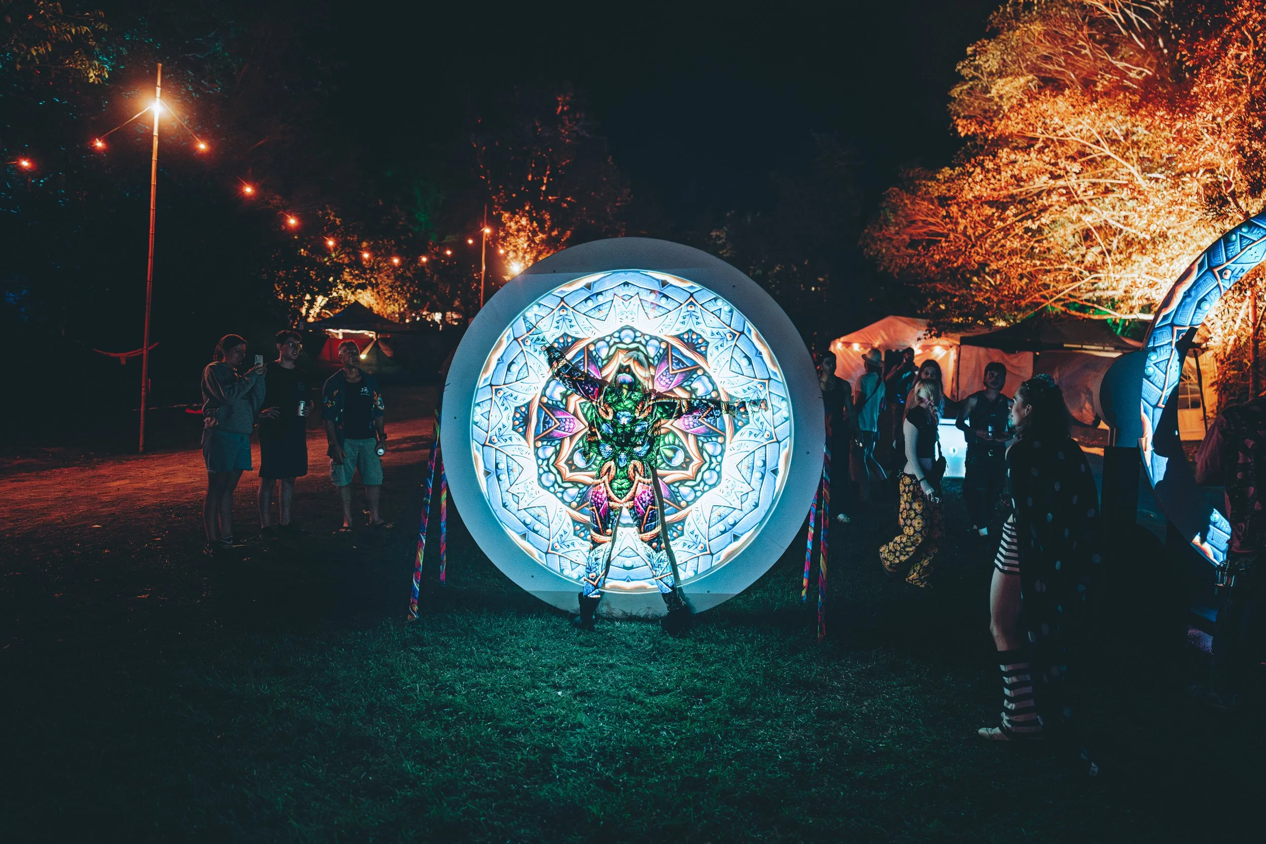Person standing in front of a large illuminated, circular, stained glass-style art piece at night, with people gathering around and trees lit with orange lights in the background.