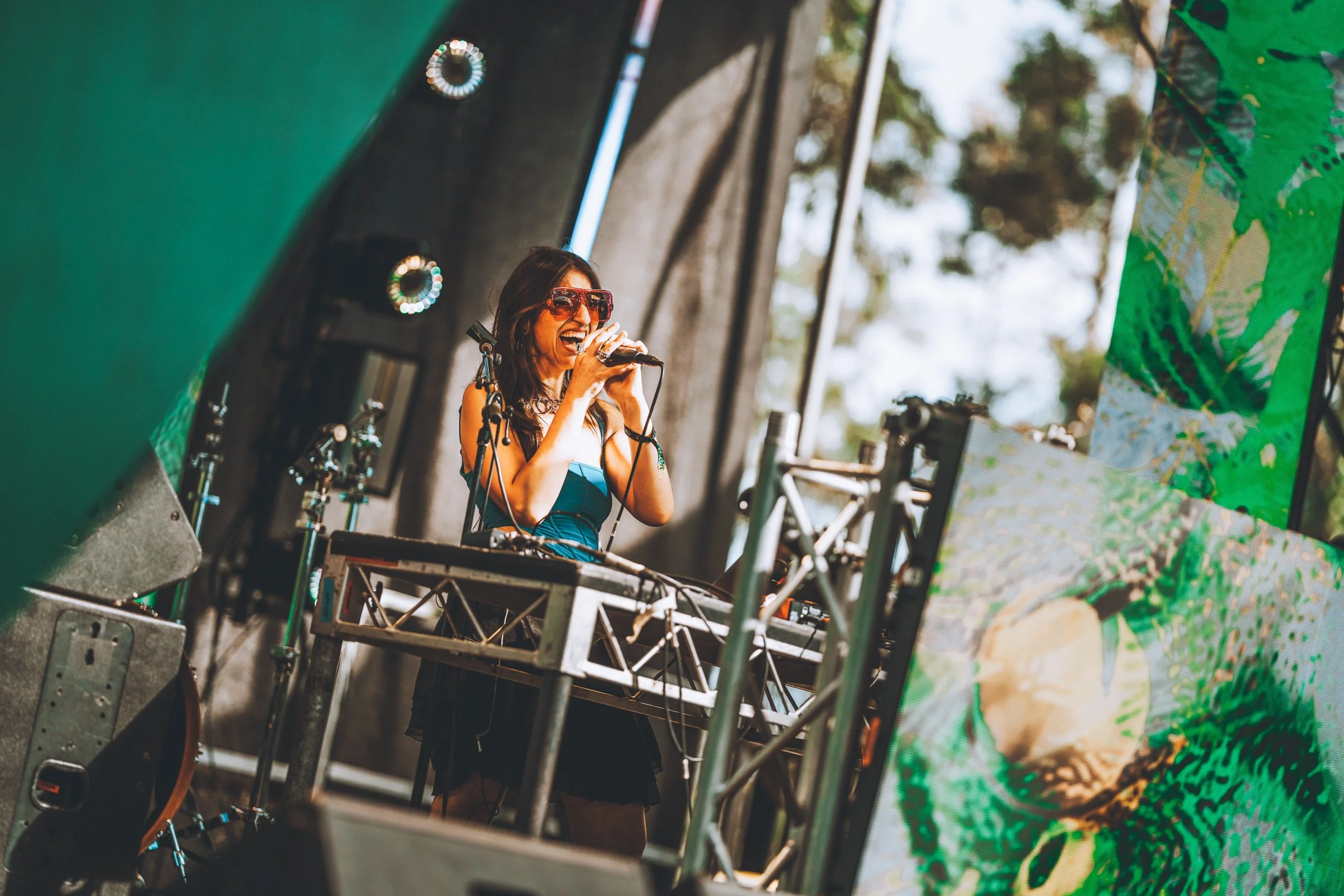 A female DJ with long dark hair, wearing sunglasses and a strapless top, is singing or speaking into a microphone on stage at an outdoor event, with trees and a digital screen showing green patterns nearby.