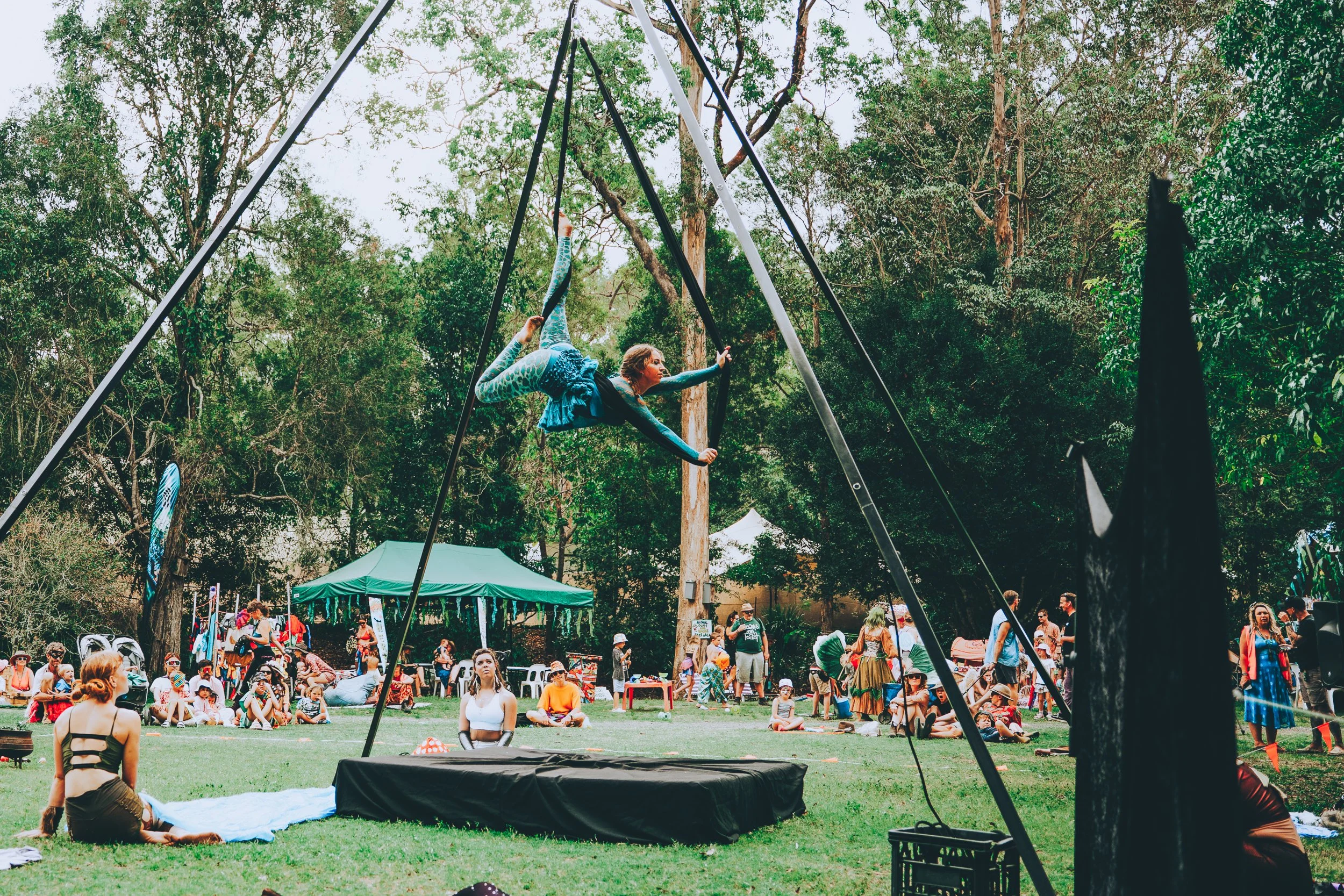Aerialist performing on a trapeze at an outdoor festival with a crowd watching, surrounded by trees and tents.