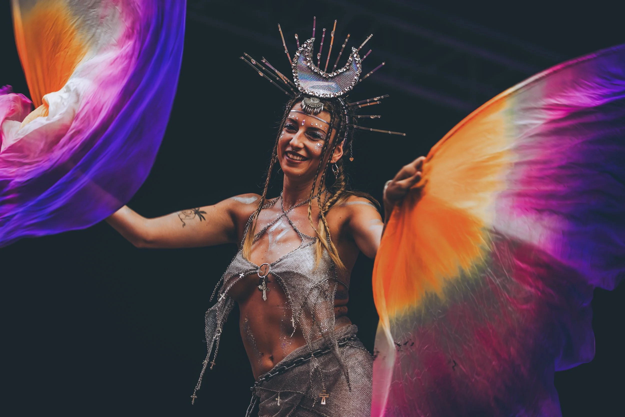 A performer wearing a crown-like headpiece and jewelry, smiling, holding colorful fabric wings, on stage with a dark background.