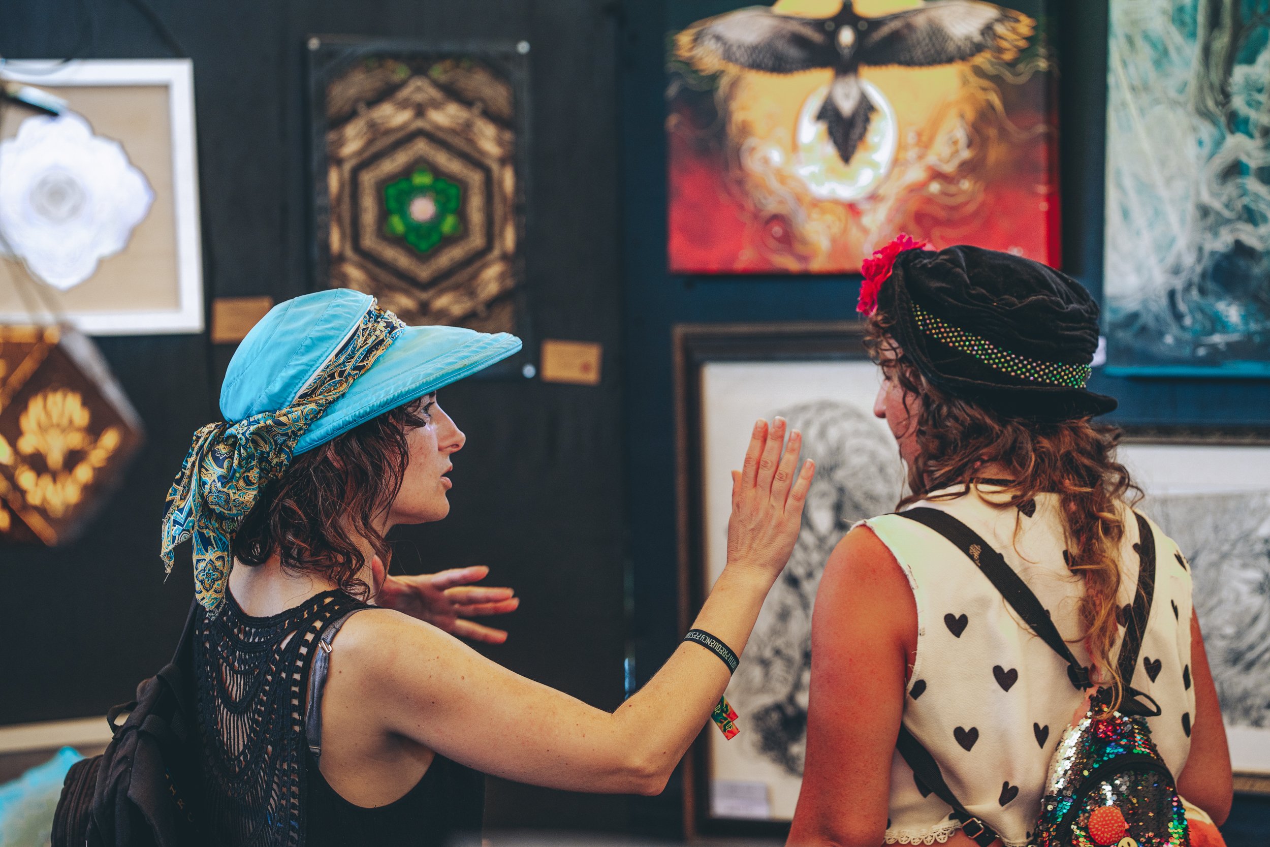 Two women having a conversation at an art gallery. One woman, wearing a blue hat with a patterned scarf, is gesturing with her hand, while the other woman, in a black hat with red flowers, listens. Artworks are displayed on the wall behind them.