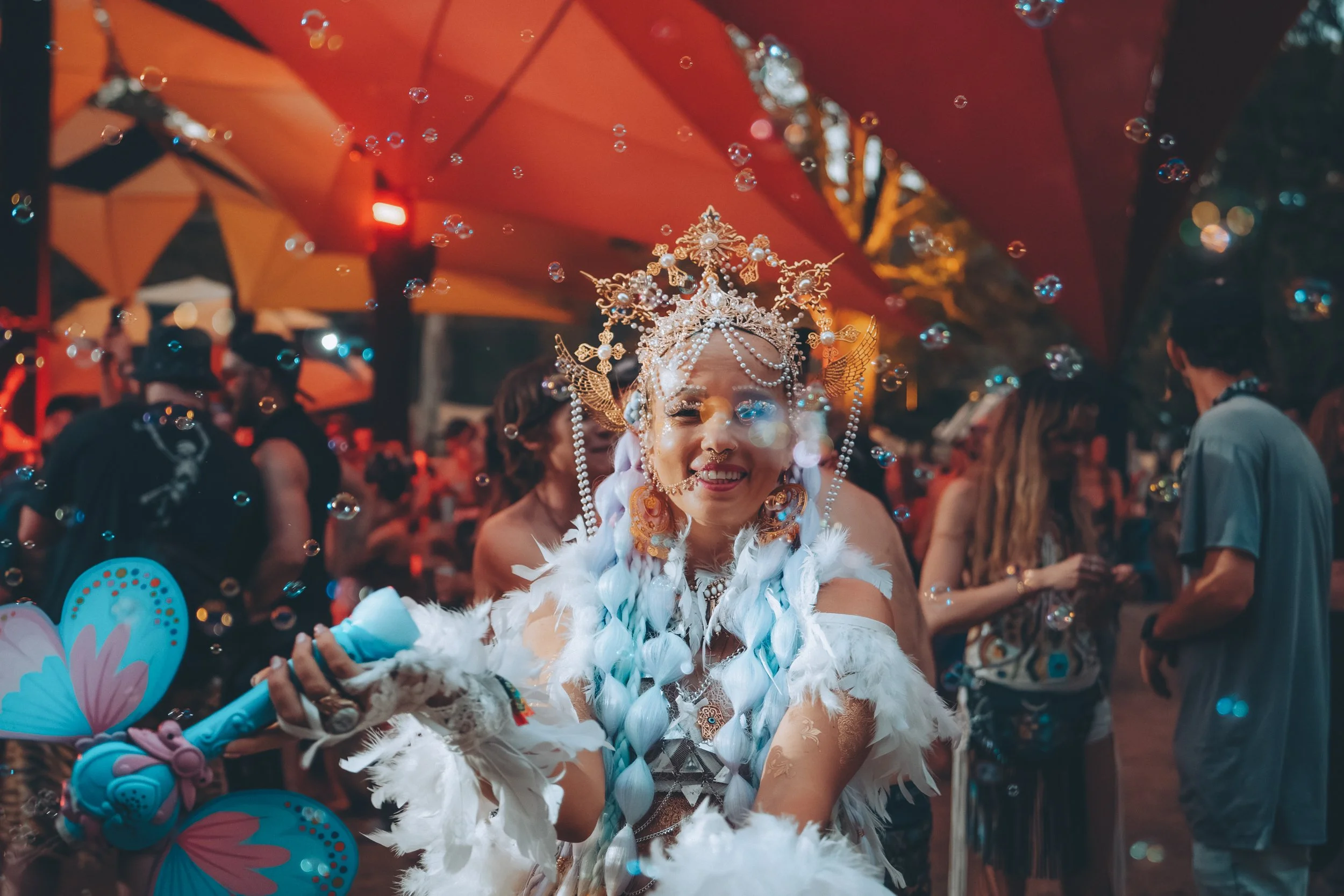 A woman dressed in a colorful, elaborate costume with pearl jewelry and feathers, holding a butterfly-shaped object, smiling at a lively outdoor event with people and umbrellas in the background.