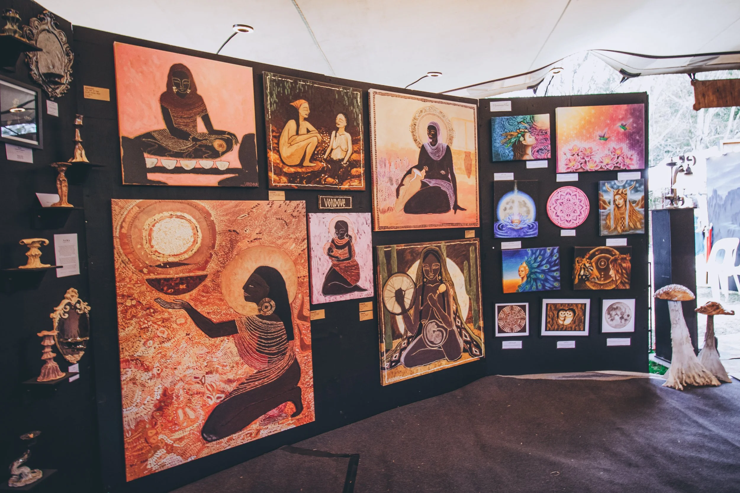 Art exhibit displaying various paintings and mixed media artworks on black display panels, featuring themes of spirituality and nature, including portraits of women, celestial symbols, and wildlife.