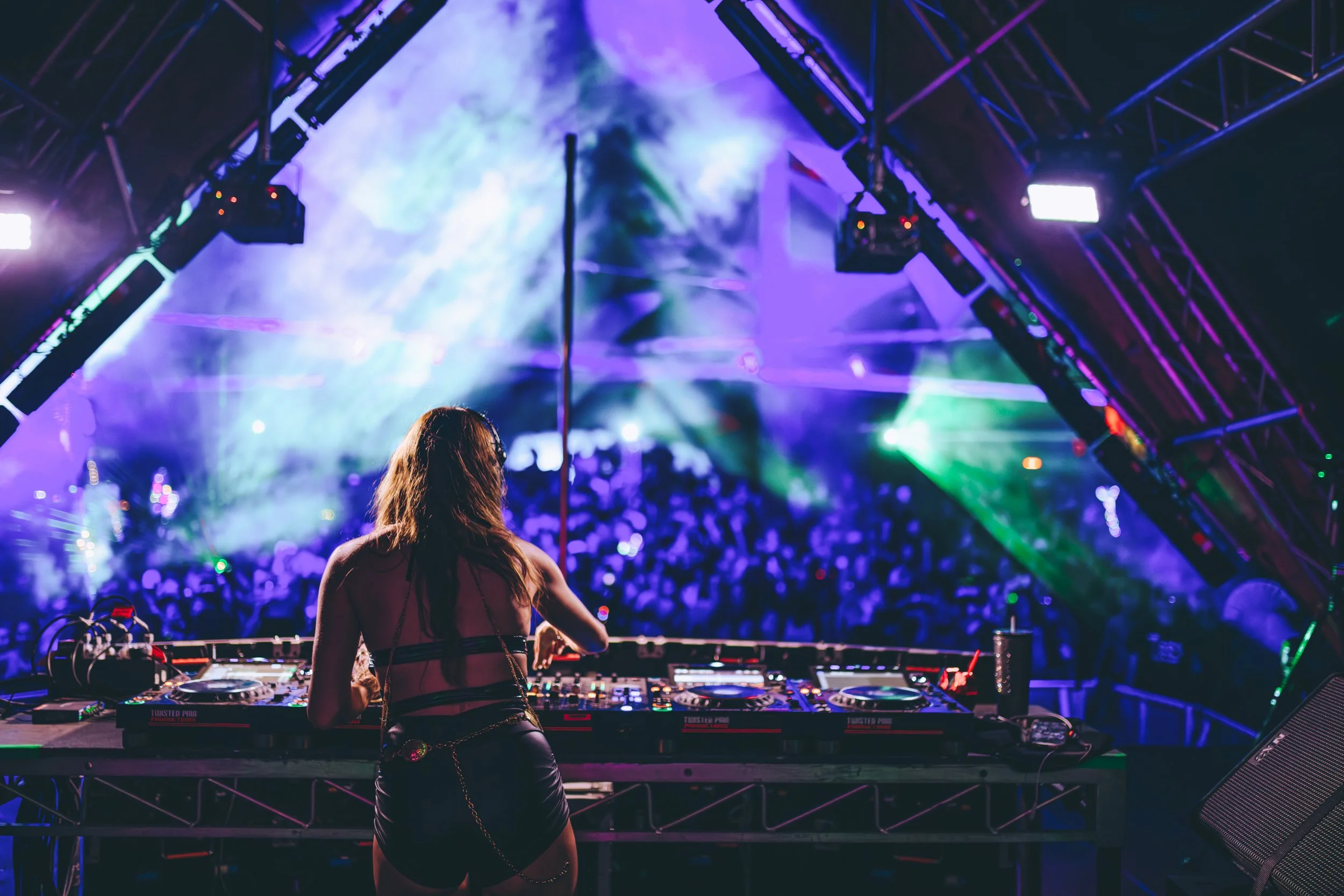 A female DJ performs on stage at a concert with colorful laser lights and a crowd in the background.