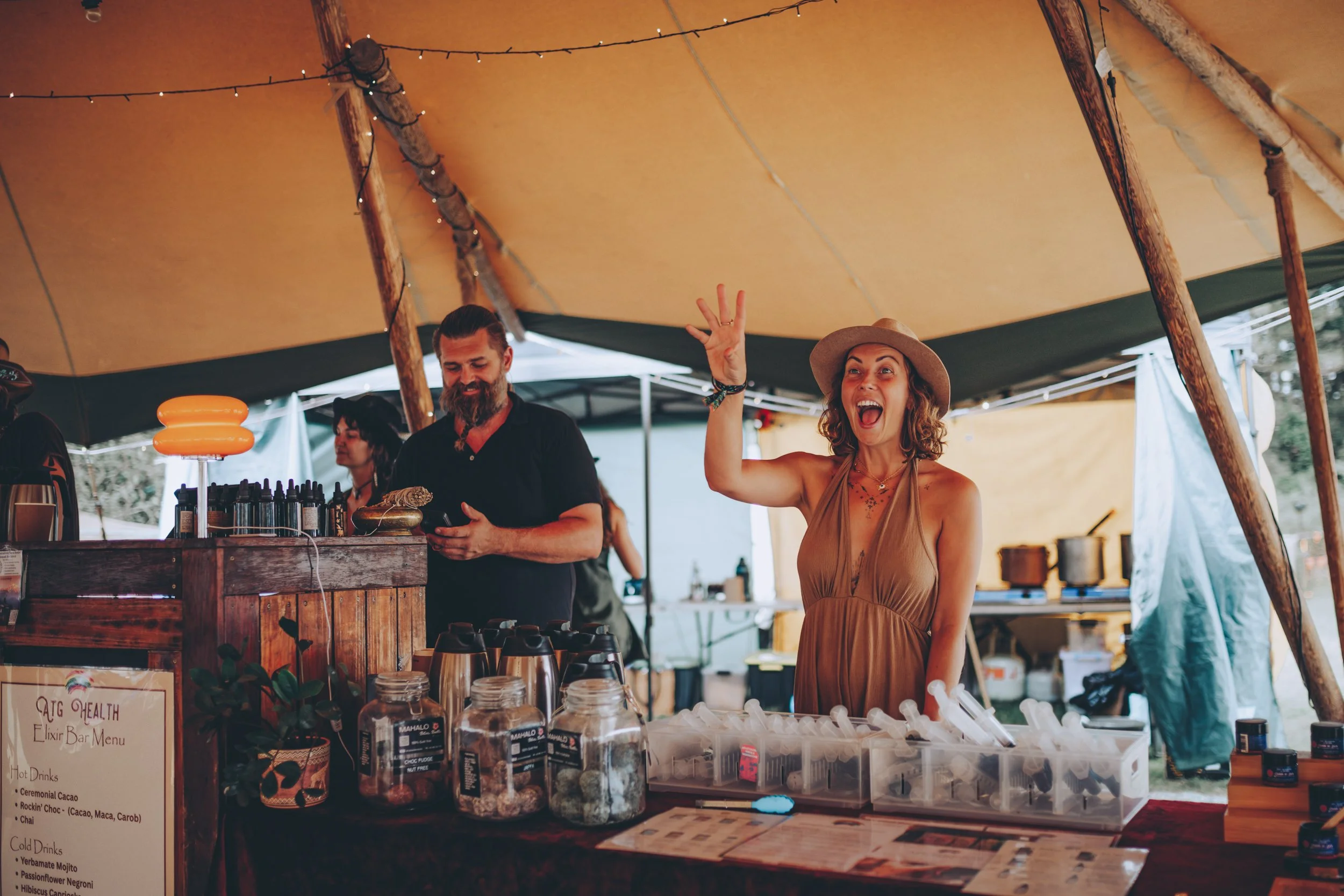 Woman in a tan dress and hat gesturing excitedly at a booth with jars and bottles, under a tent at an outdoor event, with other people and equipment visible in the background.