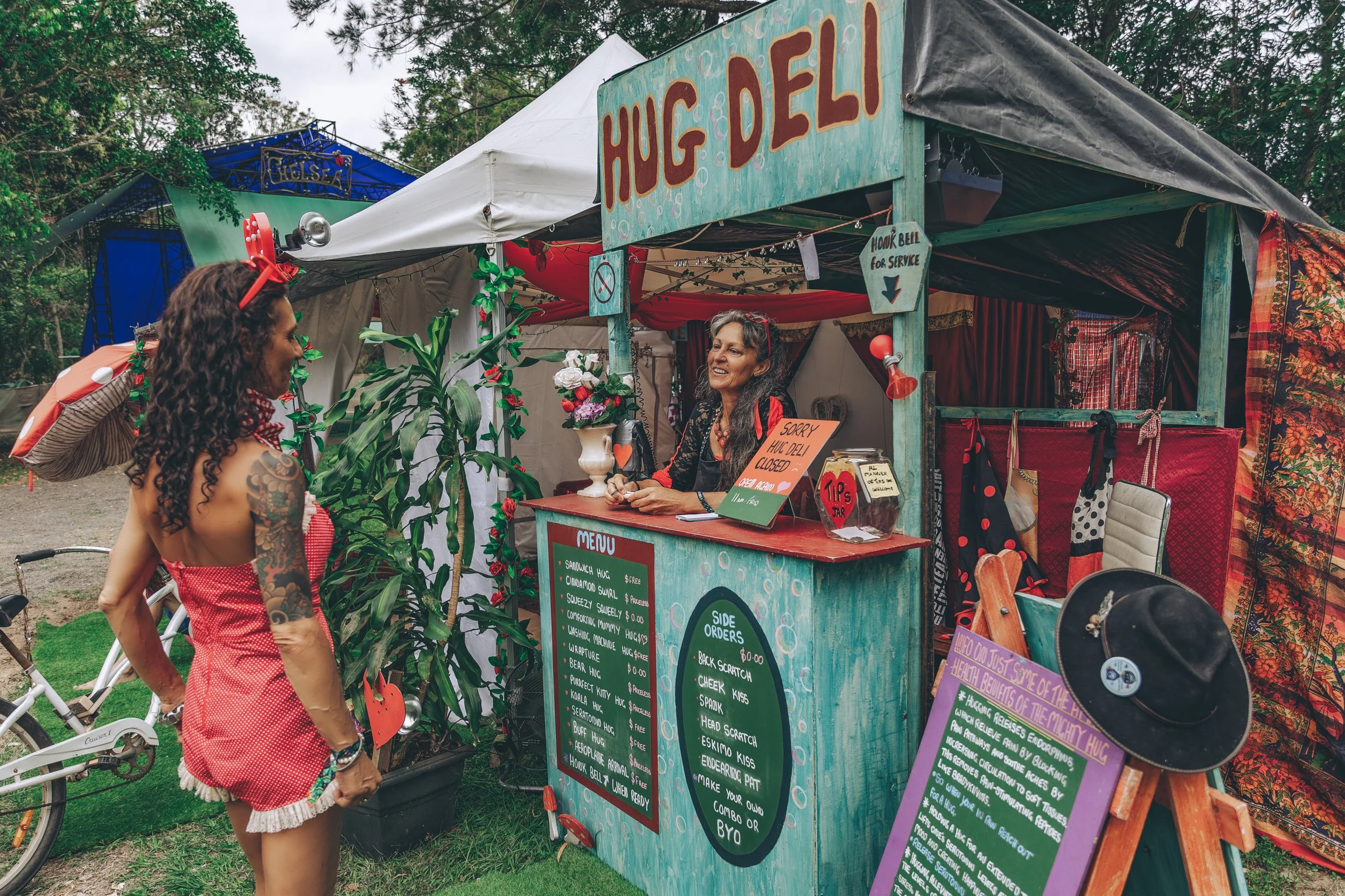 A woman with tattoos in a red dress interacts with a vendor at a small outdoor booth with a sign that says 'HUG DELI'. The booth has colorful signs with menu items, side orders, and a 'Sorry, HUG DELI closed' sign. The vendor woman is smiling behind 