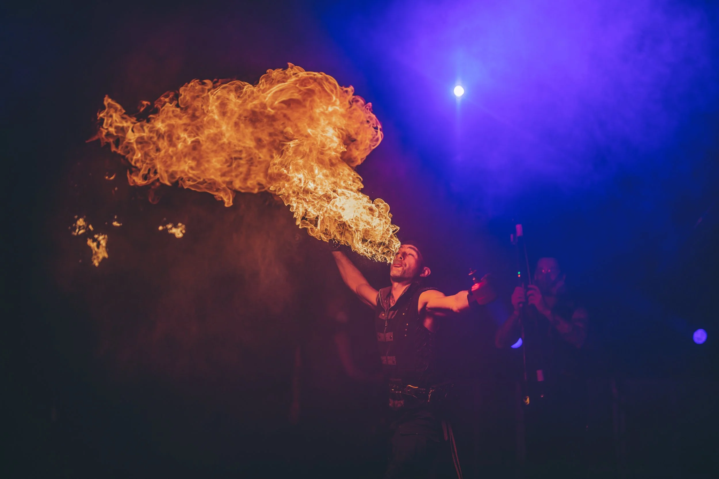 A fire performer spewing fire into the air during a night event, with purple lighting in the background.
