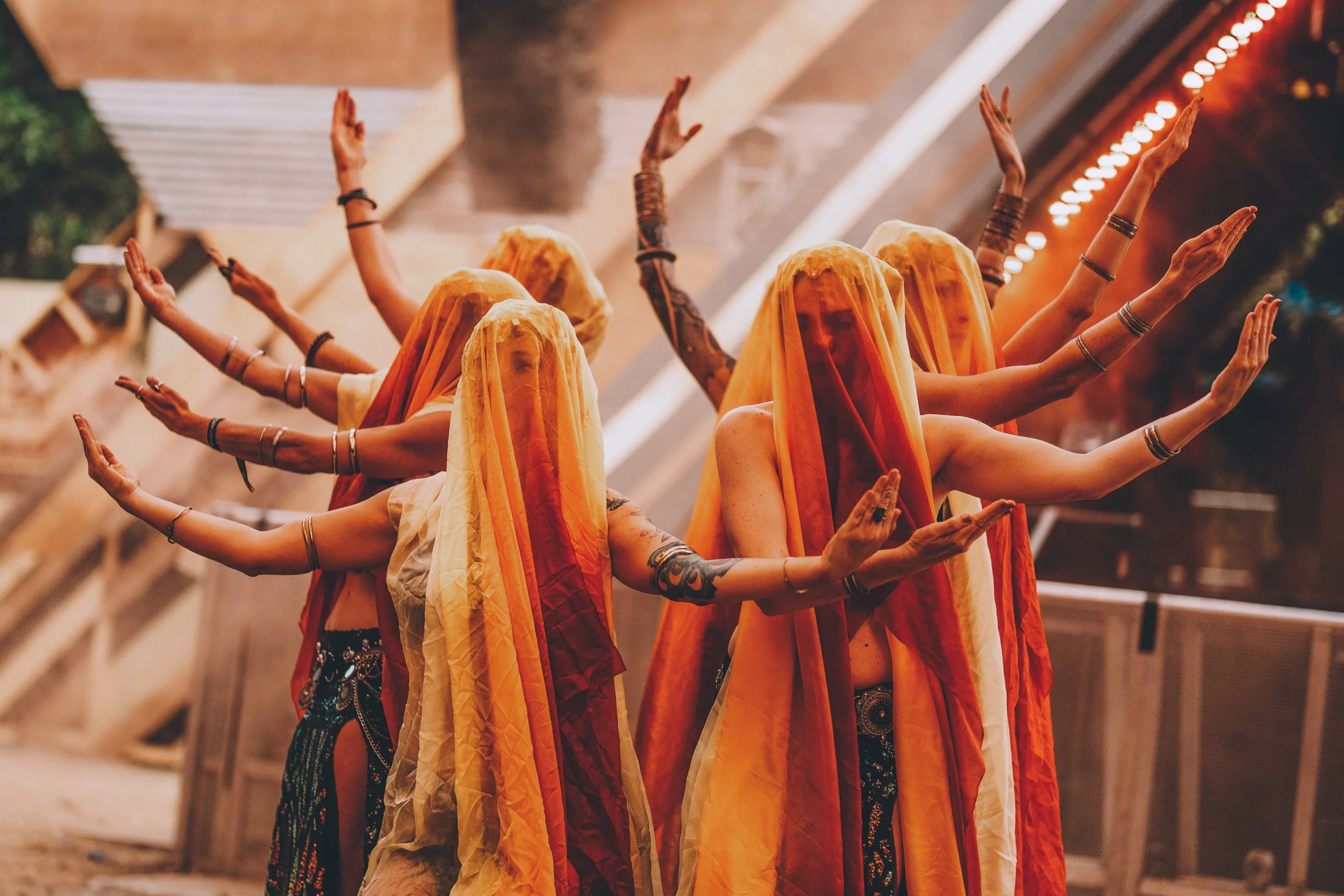 Women dressed in flowing orange and yellow garments with veils covering their faces, performing a traditional dance with arms raised and joined, outdoors at night.