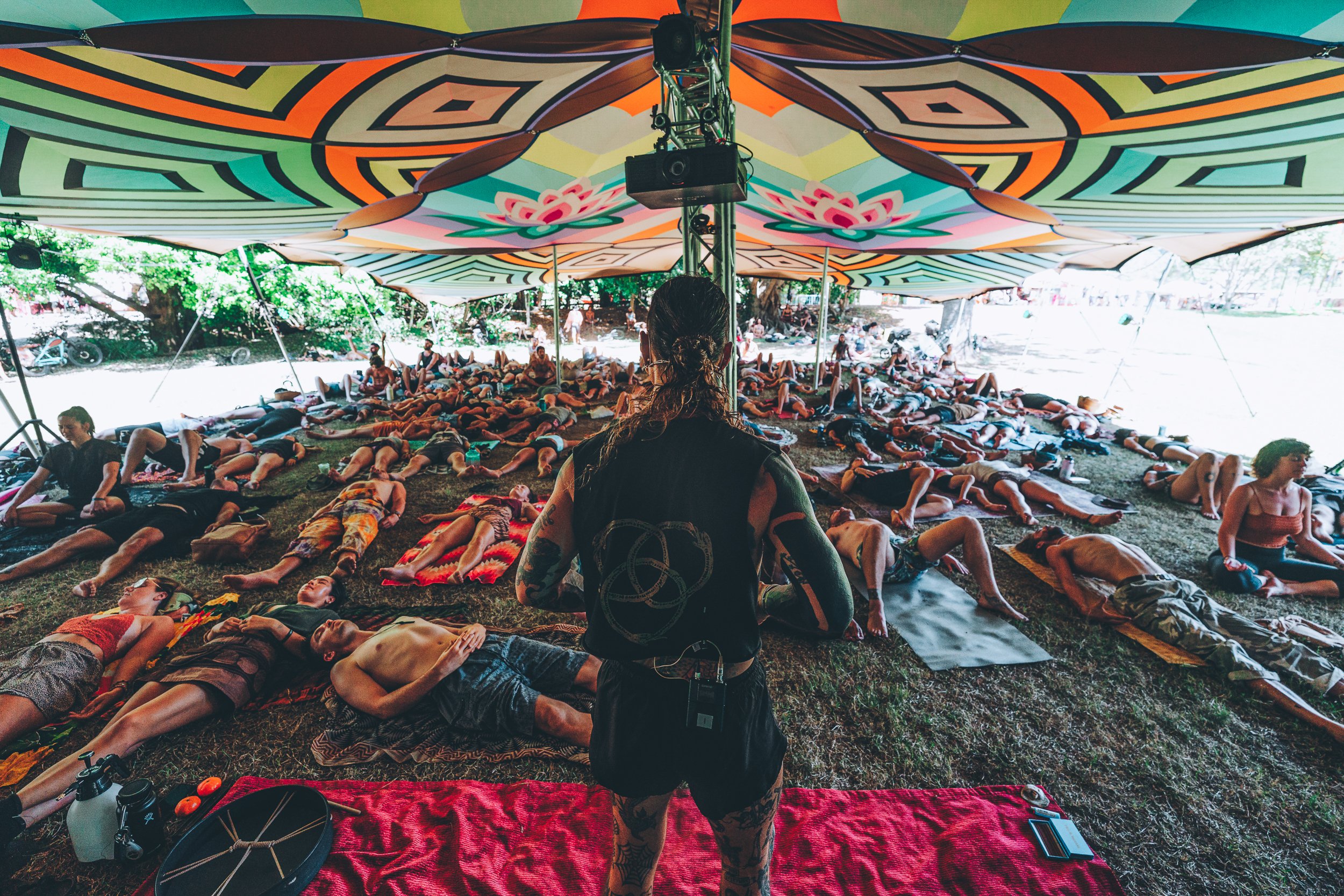 A large group of people participating in a yoga class under a colorful tent outdoors, with the instructor standing in front of them.