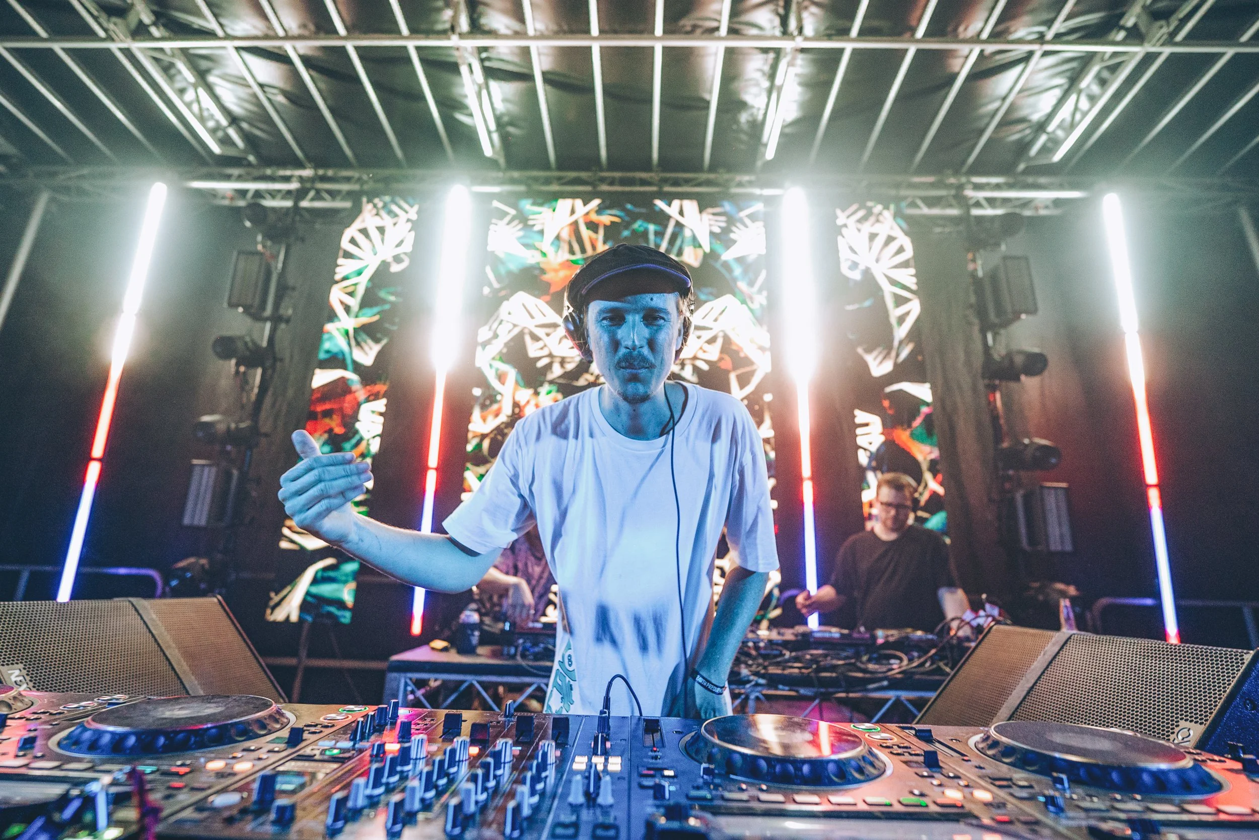 DJ performing at a concert with electronic equipment in front and colorful visuals behind on a large screen.