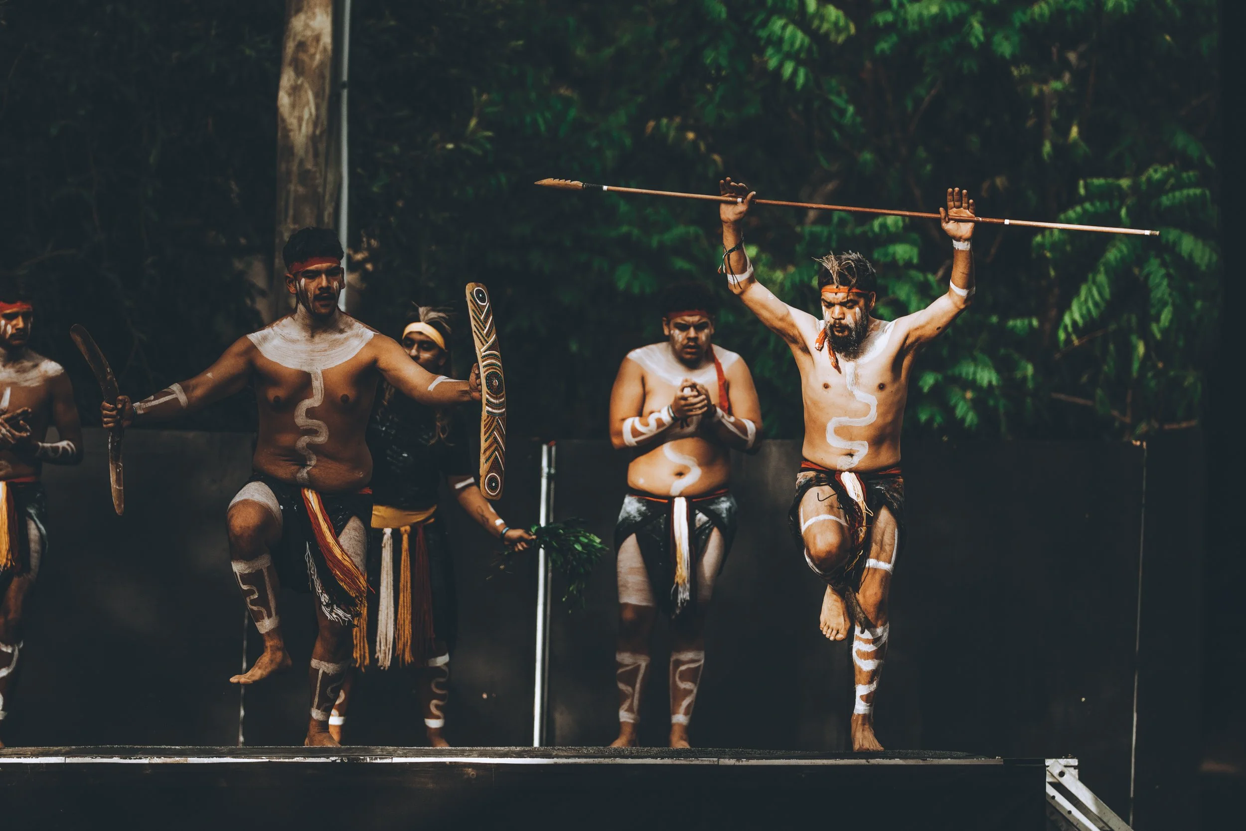 Indigenous men performing a traditional dance on an outdoor stage, dressed in traditional attire with body paint, holding shields and spears, with a forest background.