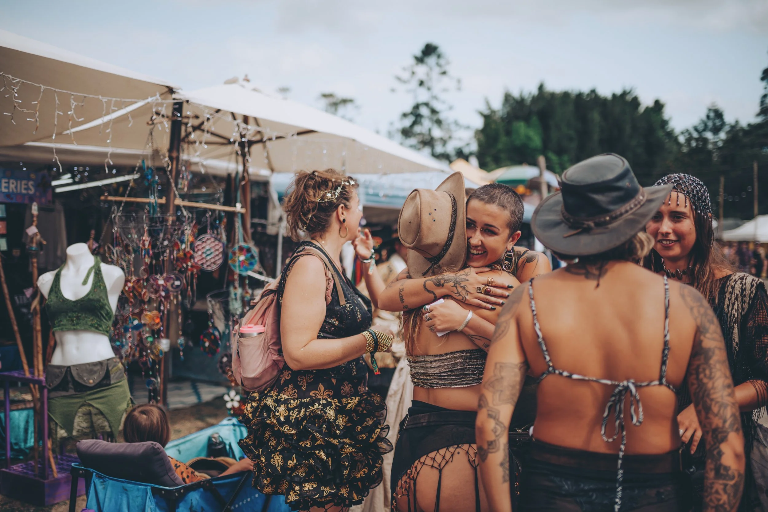 Group of women hugging and smiling at a street market, with clothing and jewelry stalls in the background.
