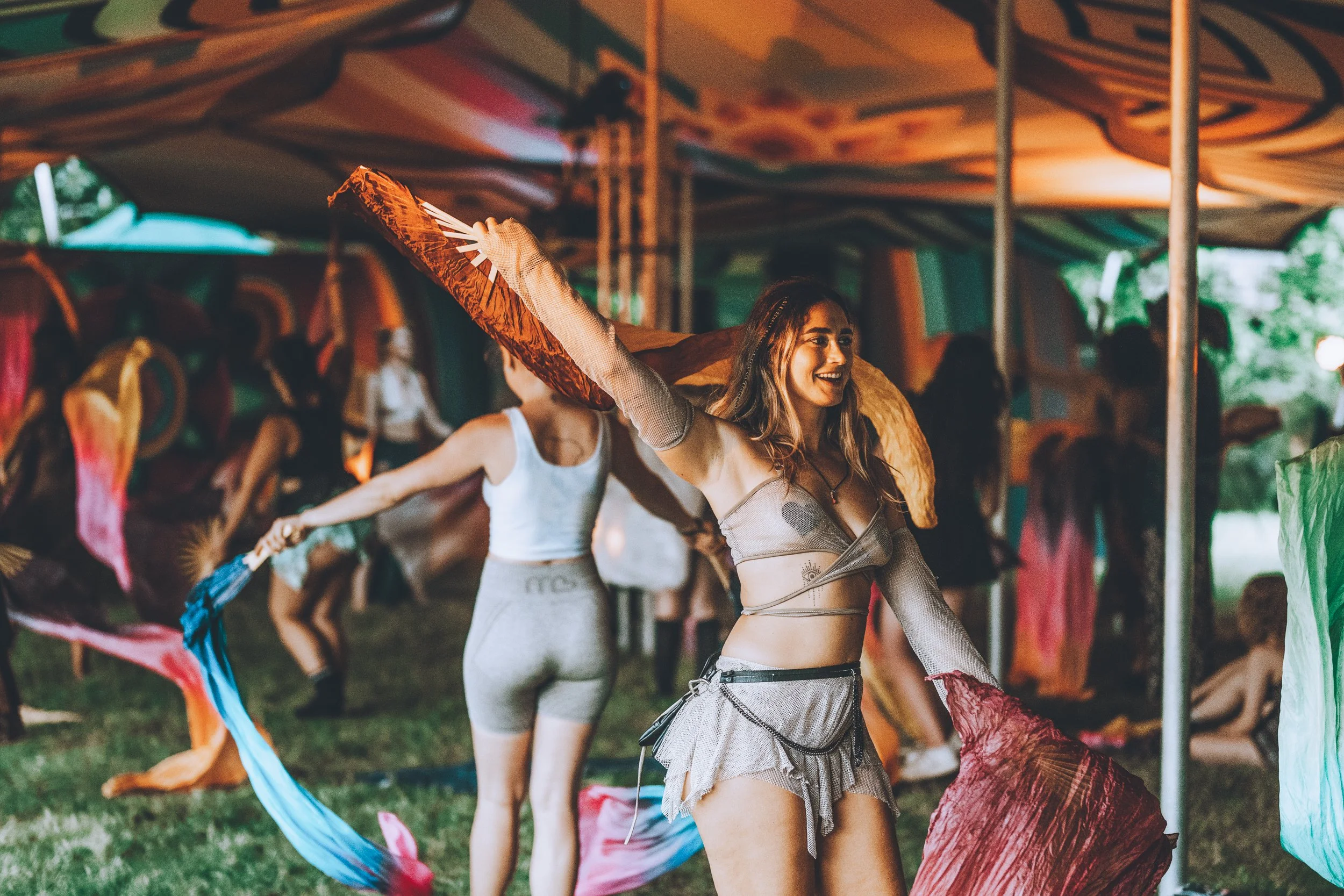 Women dancing with colorful fabric flags at an outdoor festival or event under a tent.