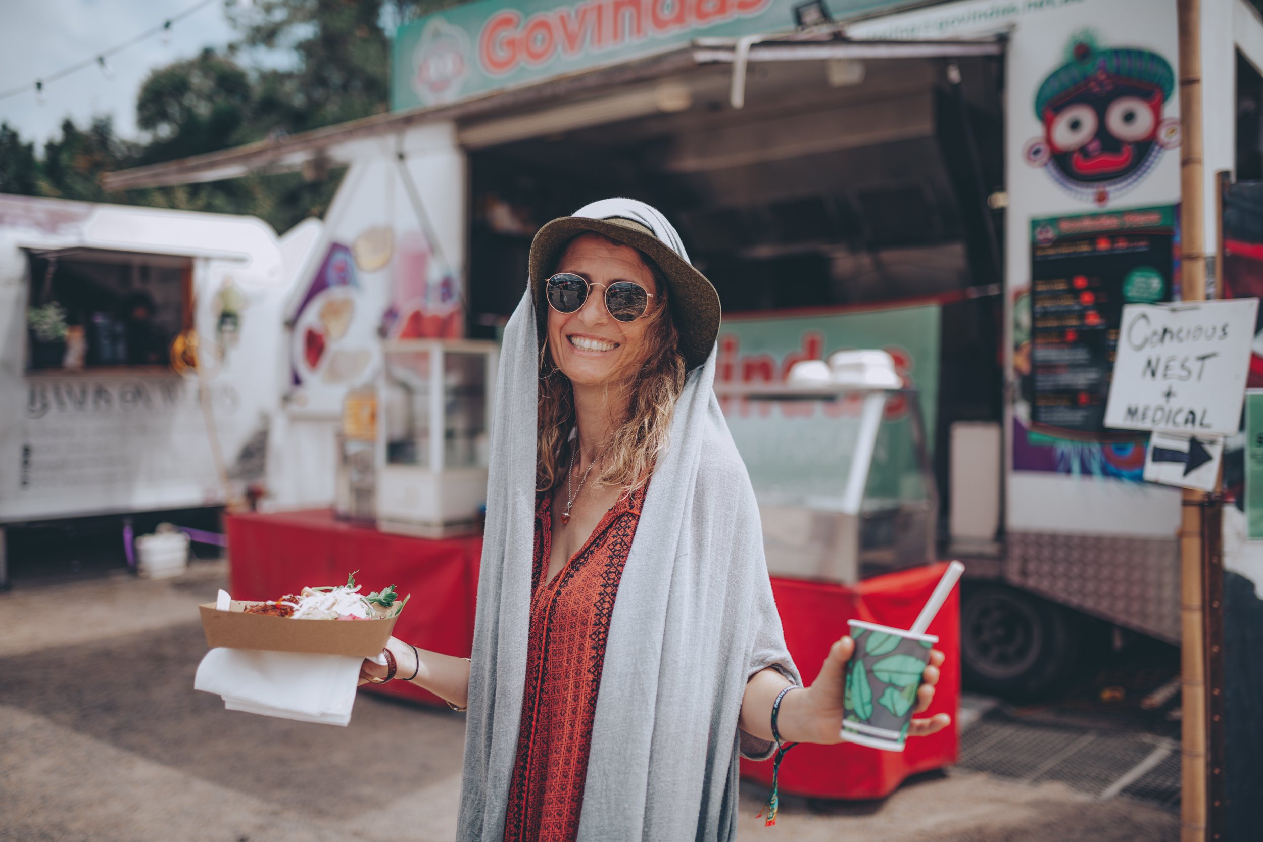 A woman smiling and holding a tray of food in one hand and a beverage in the other at an outdoor fair or market, wearing sunglasses, a wide-brimmed hat, a red patterned dress, and a gray shawl.