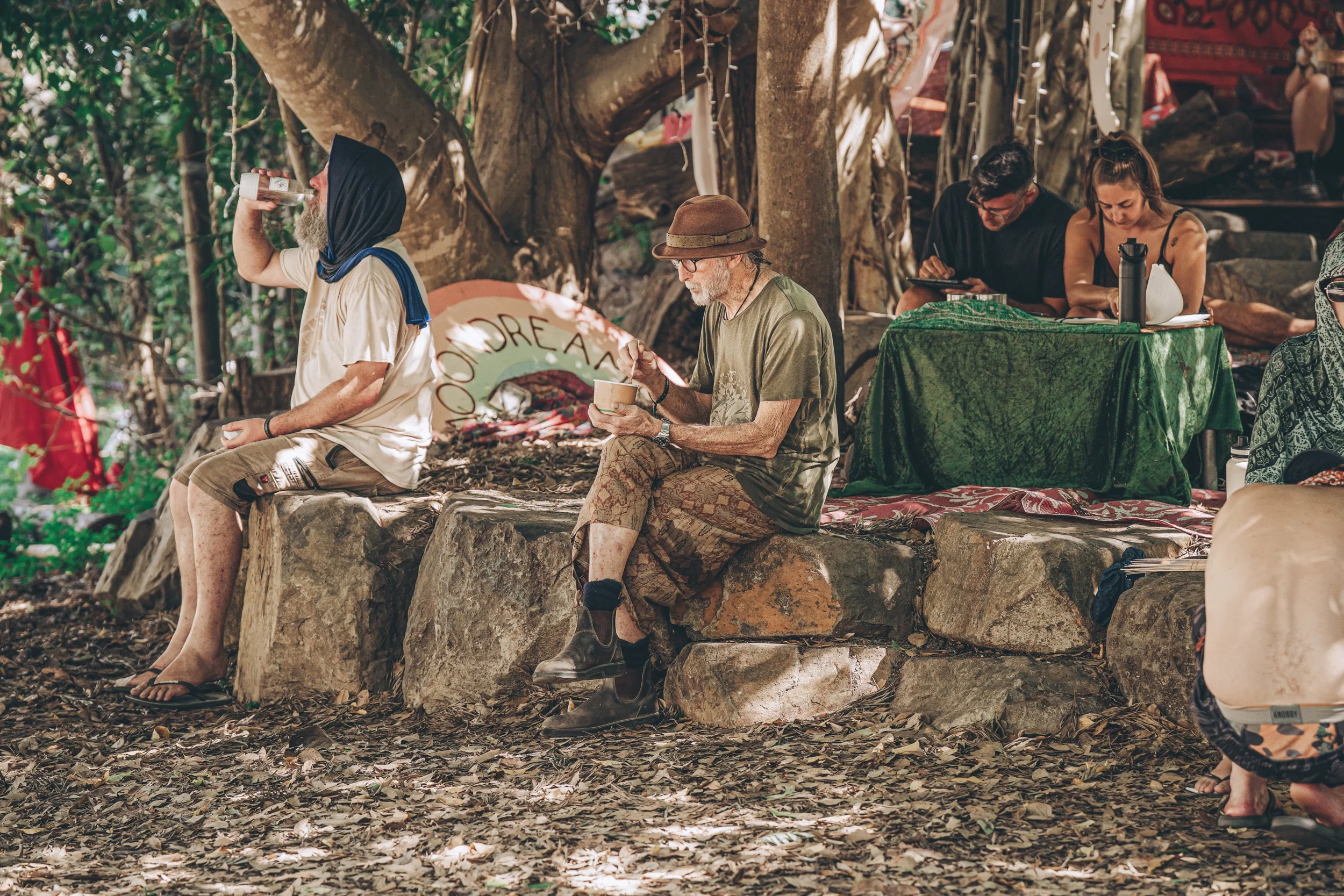 People sitting on rocks and at a table outdoors under trees, some drinking or writing, in a natural, shaded setting.