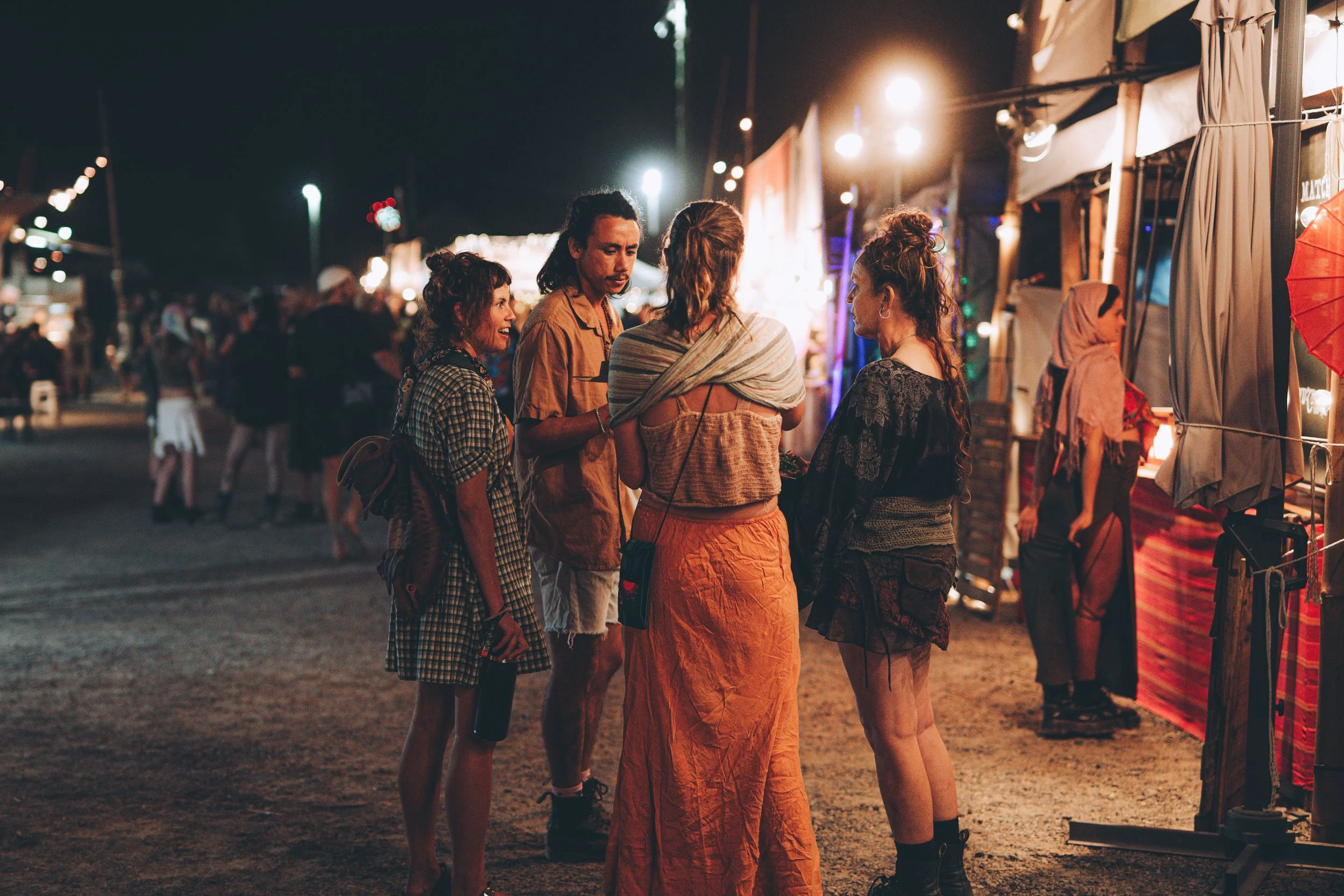 Group of five young adults talking at night at outdoor fair or carnival with food and craft stalls.