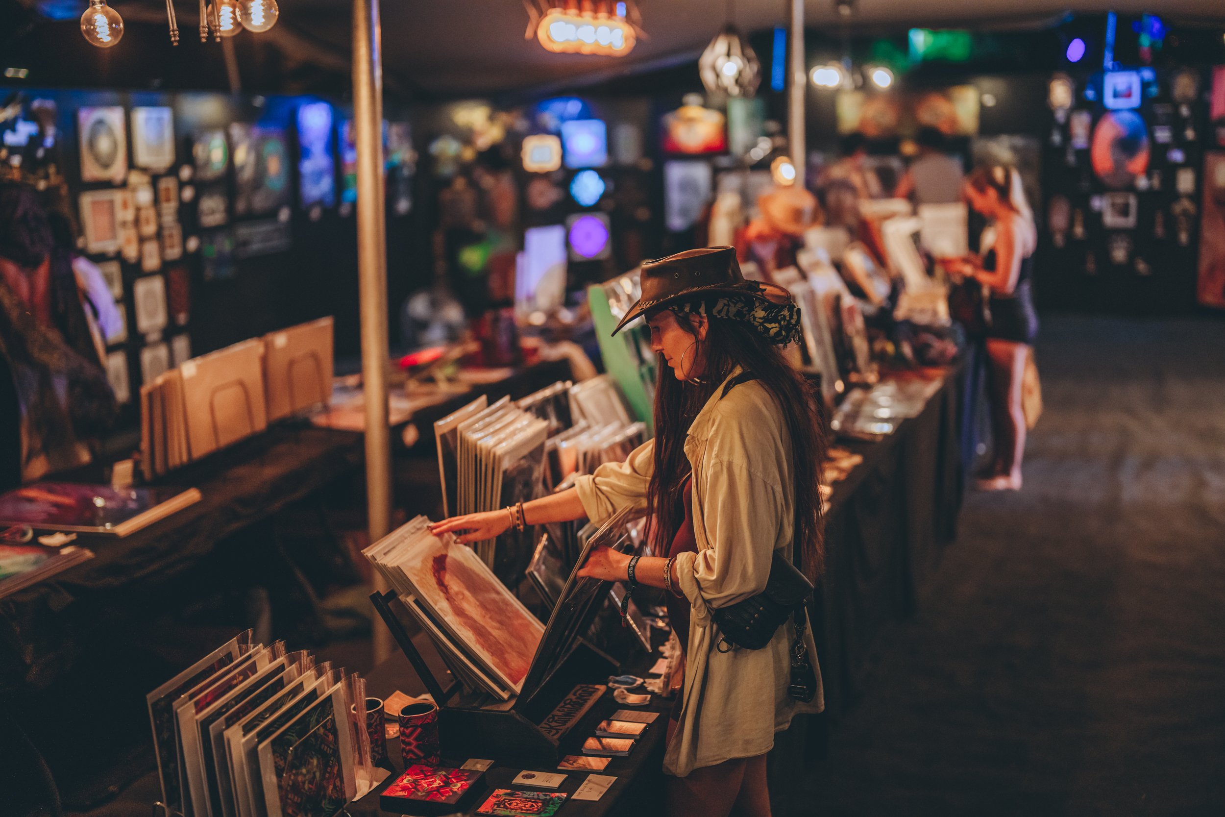 A woman in a cowboy hat browsing through artwork or prints at an art fair or gallery, with other people shopping and artworks displayed in the background.
