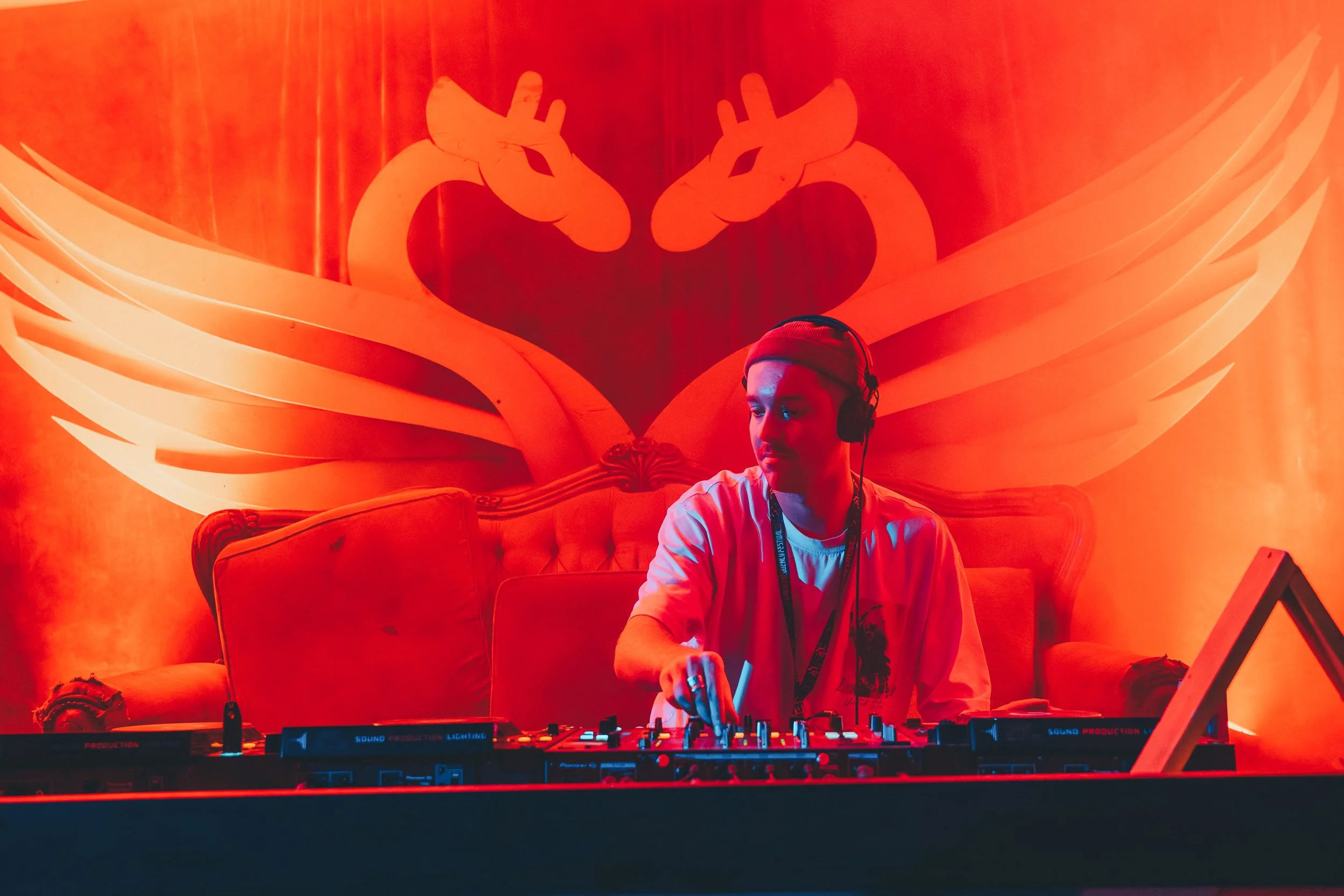 A DJ wearing a red beanie and headphones is performing at a club with a large illuminated backdrop of a stylized double-headed dragon with wings in the background.