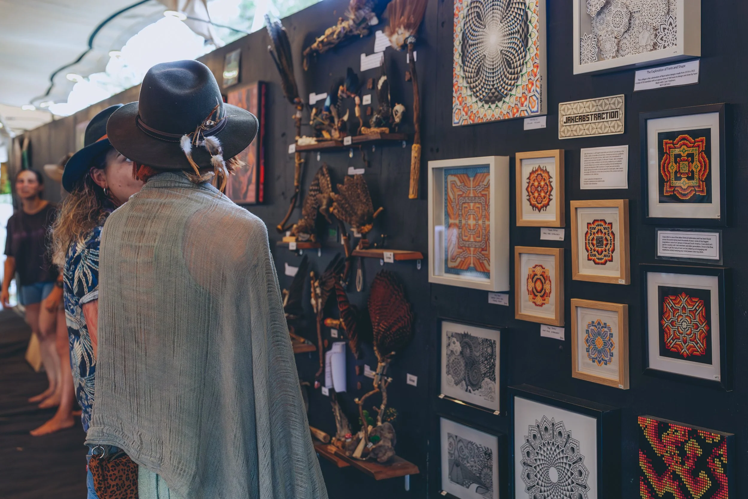 People at an art exhibit, looking at framed artwork and crafts on a dark wall. The artwork includes colorful geometric patterns, black-and-white designs, and 3D textured pieces, with some sculptures and woven items displayed below.