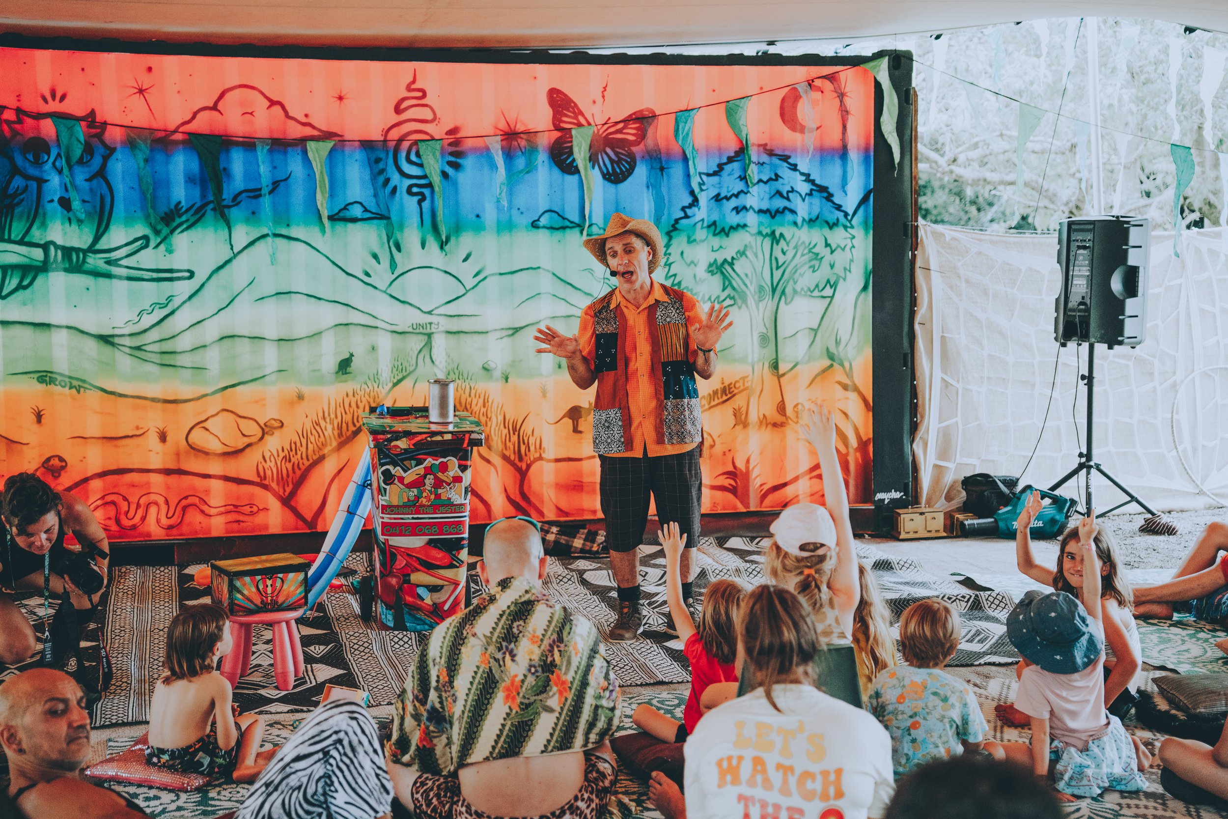 A clown performing a show on a stage with a colorful backdrop, entertaining a group of children and adults sitting on the floor.