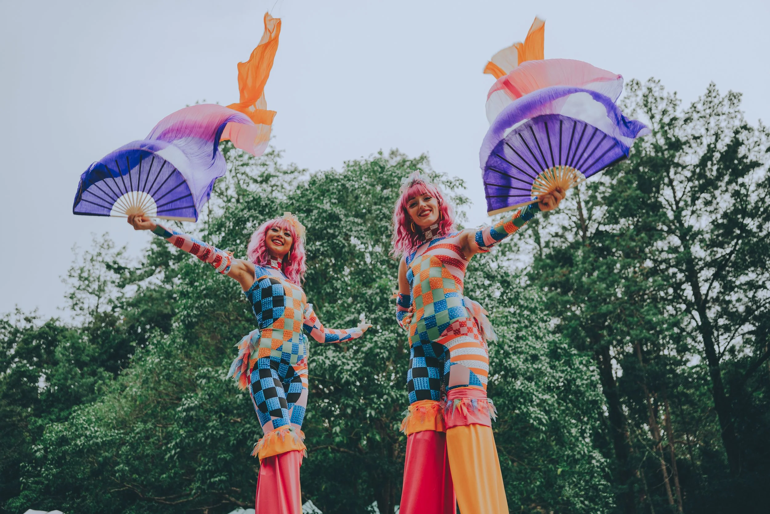 Two performers with pink hair in colorful patchwork costumes on stilts, holding purple and orange fans, smiling in a park with green trees.