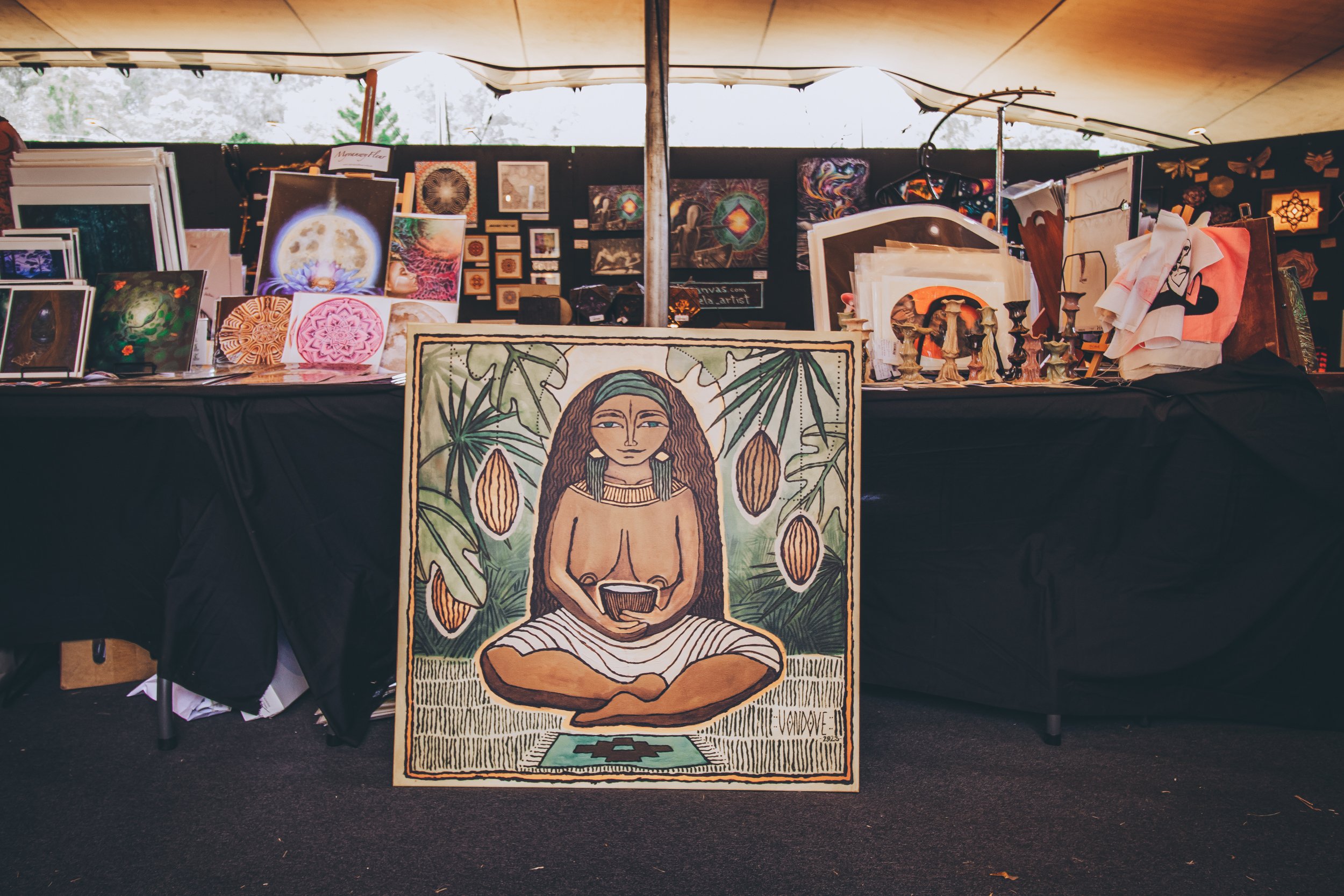 An art booth display featuring various paintings and artworks, with a prominent painting of a woman with long hair sitting cross-legged, holding a bowl, surrounded by lush green leaves and cacao pods.