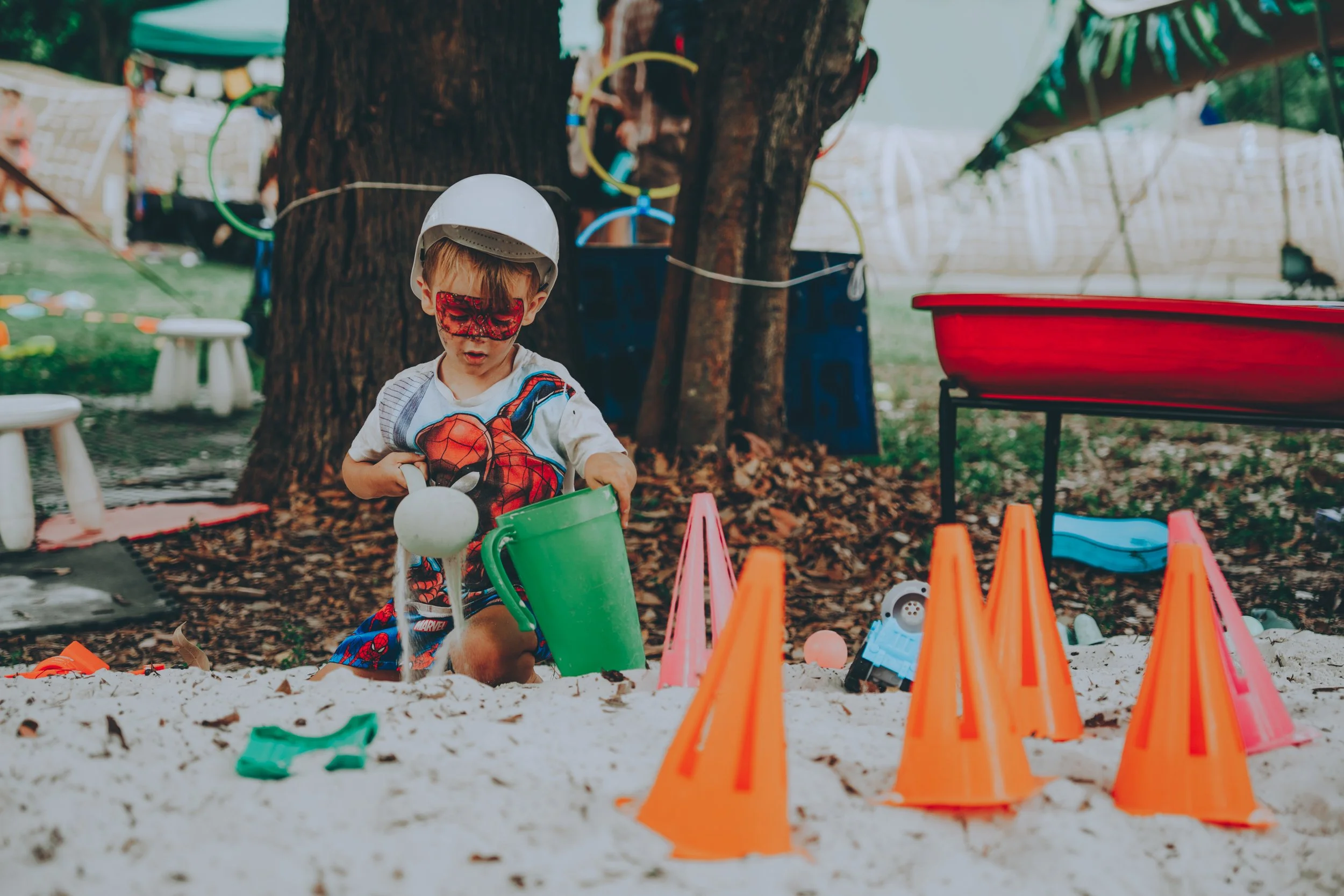 A young boy wearing a Spider-Man shirt and a white helmet playing in a sandbox with toy shovels and cones, surrounded by trees and outdoor play structures.