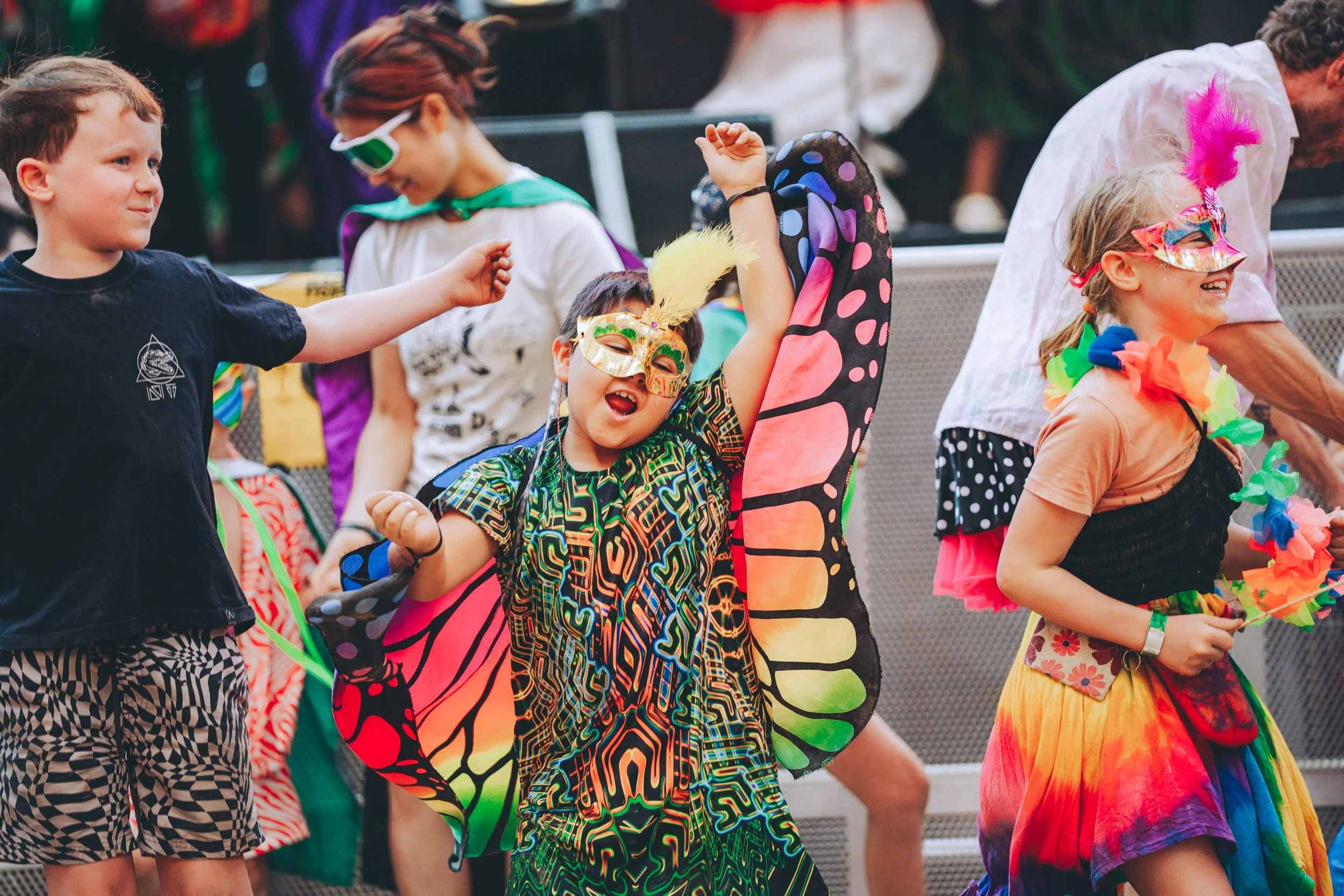 Children at a festive outdoor event wearing colorful costumes and masks, some dancing and smiling.
