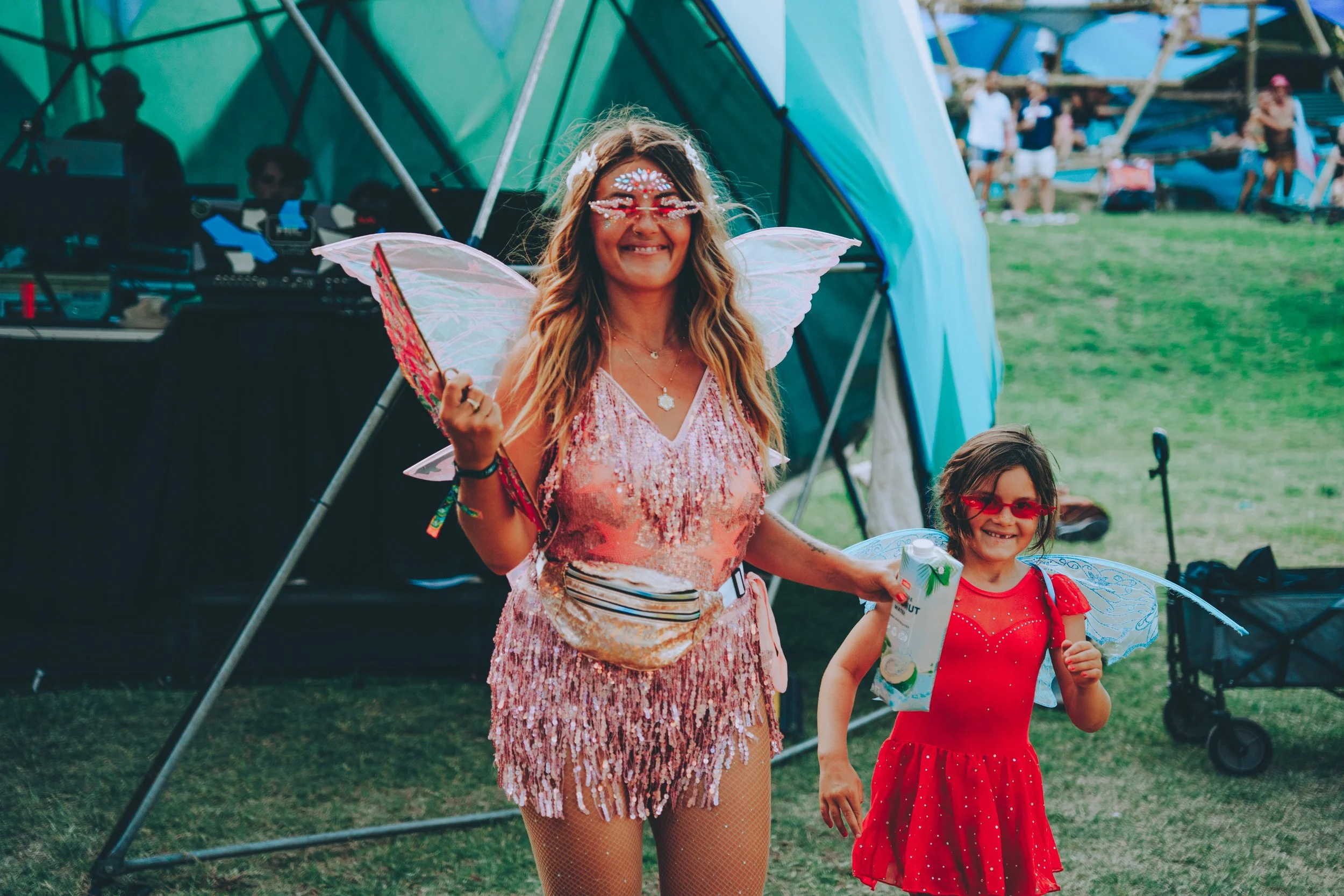 A woman dressed as a fairy with pink sequin dress, fairy wings, and face decorations, smiling and holding a toy wand. A young girl in a red dress with fairy wings and glasses stands beside her, smiling, holding a snack, with a music setup and people 
