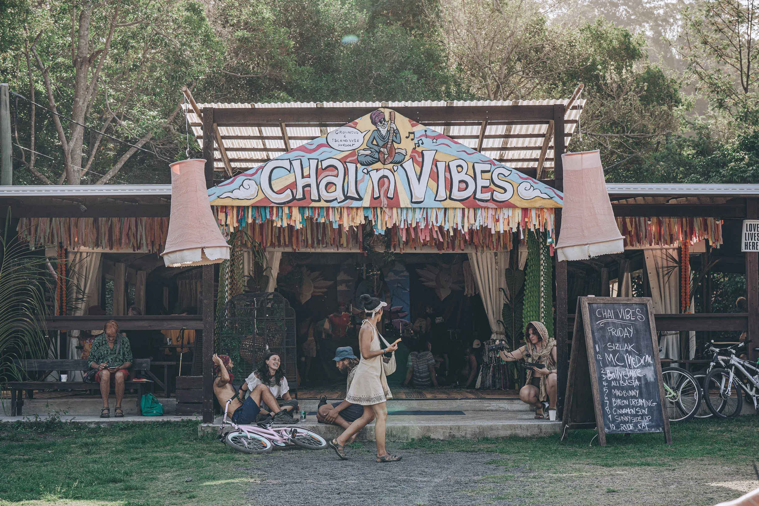 A lively outdoor scene at a venue called Chai n Vibes with a colorful sign and decorations, several people, a pink bicycle, and a chalkboard listing Friday music acts.